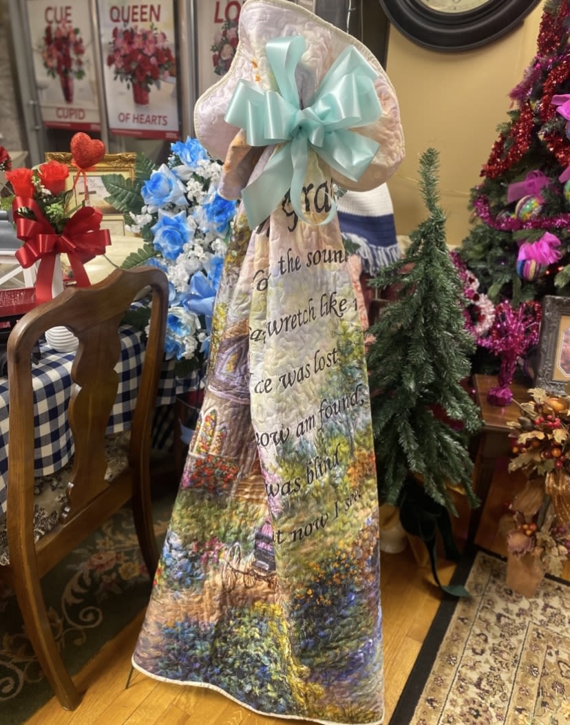 Amazing Grace Quilted Throw With a Church Scene - A gentle, commemorative quilt draped with a soft aqua satin bow-this memorial-style throw blends floral imagery and a heartfelt script to create a sympathy gift that comforts. As a remembrance gift for a funeral or to mark an anniversary of loss, the mixed-color garden scene (soft blues, pinks, and whites) reads like a keepsake someone will reach for again and again to feel remembered.  The fabric features a printed floral landscape with roses and other blossoms arranged around a central block of script text, giving the piece a handcrafted, reflective look. Tied at the top with a large aqua satin ribbon, the throw is presented upright so the sentiment is easily read; the pastel palette and quilted texture add warmth and a soft tactile comfort. Displayed here with tasteful shop arrangements, it's designed to pair well with sympathy roses or a simple framed sign to complete a memorial tribute.  For someone grieving, this throw offers a quiet way to say, "I'm thinking of you." It's sized to fold into a lap blanket or drape over a favorite chair as a lasting keepsake-an intimate gift that brings comfort long after the service. Order with same-day pickup or local delivery where available to have it arrive beside flowers or other sympathy pieces.