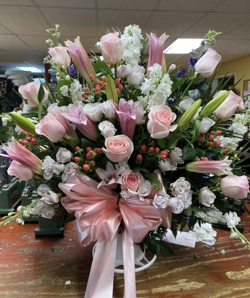 Pretty in Pink Funeral Floor Basket  - A lavish cascade of soft pink roses and open pink lilies creates an elegant sympathy and funeral arrangement. This tall, formal flower arrangement presents a comforting, room-filling silhouette: the recipient will feel gently seen by its graceful blend of pinks, creamy whites, and subtle green and purple accents.  At the center lies a generous mound of pink and white roses layered with white stock, pink lilies, spray roses and delicate berry accents for texture. The composition radiates outward from a substantial base, incorporating touches of carnations, alstroemeria, snapdragons, stock and Bells of Ireland to add height and movement. Finished with a wide pink satin ribbon and displayed on a white pedestal stand, the piece reads as a polished tribute with a refined, classic palette.  Designed for formal display on a floor stand or large table, this substantial arrangement brings calming presence to memorial services while remaining appropriate for celebratory moments that call for dignity and warmth.