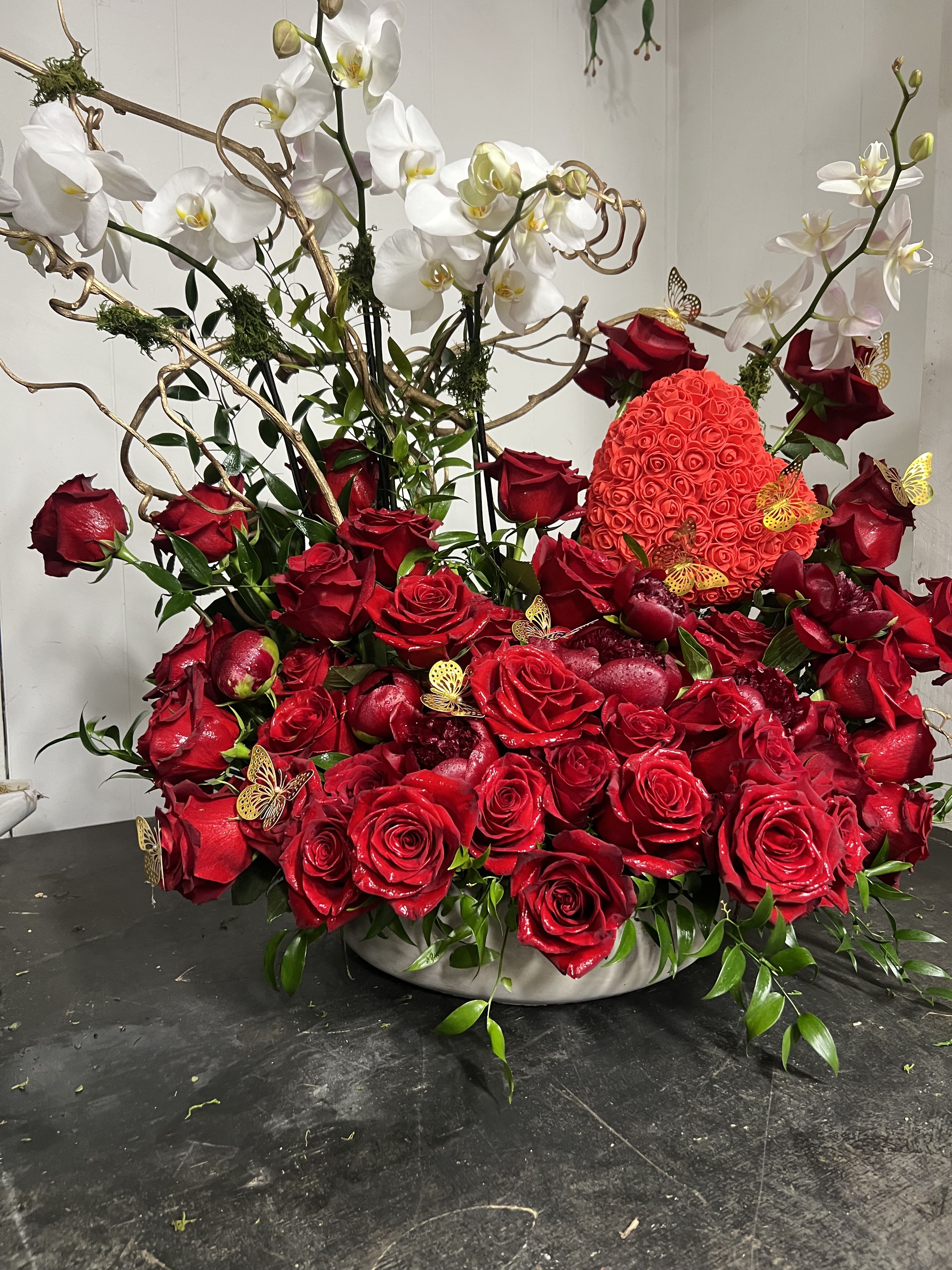 Amor Eterno  - A lush mound of velvety red roses anchors this Amor Eterno flower arrangement, while sweeping white Phalaenopsis orchids arch above for a sculptural, high-contrast statement-an elegant choice for an anniversary, Valentine's Day, or any love-and-romance moment when you want the person you're thinking of to feel cherished. The dramatic red-and-white palette also works beautifully for formal celebrations, holiday centerpieces, or to mark a milestone like a retirement, graduation, or birthday.  Set in a low ceramic container, the design combines a dense, rounded base of roses with airy orchid stems and curly branch accents that add vertical motion and texture. Small decorative butterfly picks give a celebratory, whimsical note without overwhelming the classic color story. The overall size reads as generously abundant yet refined, making it suitable for a mantle, dining table, or reception desk.  Perfect for sending as a romantic gesture, a heartfelt thank-you, or a sophisticated get-well or sympathy tribute, Amor Eterno translates across seasons-Autumn and Christmas receptions, Spring and Summer weddings, or quieter moments of "thinking of you." Place in fresh water and keep away from direct heat; orchids and roses will reward gentle care so the recipient can enjoy the arrangement for days.