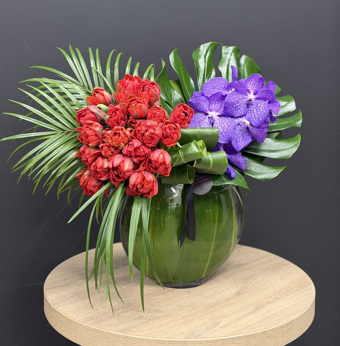 Marrakech  - A bold, sculptural arrangement inspired by the vibrant energy of Marrakech. Rich red double tulips meet striking purple Vanda orchids, layered with glossy tropical greens and rhythmic leaf work. Designed in a modern moon glass vase lined with lush foliage, this piece brings a luxurious, vacation-like statement to any space. Perfect as a statement centerpiece or a signature gift, Marrakech delivers color, texture, and refined style in every stem. Height: 12-14 inches Width: 14–16 inches