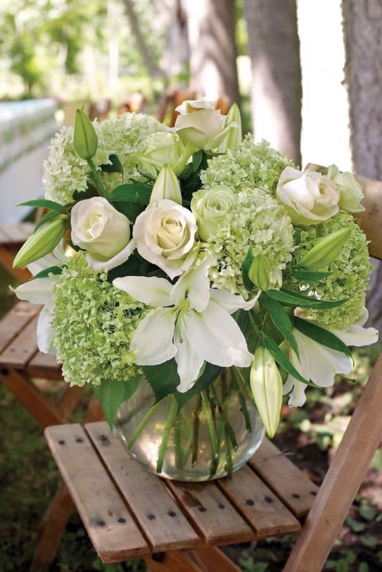 White lilies, roses, and green hydrangeas in a clear glass vase on a wooden stool outdoors