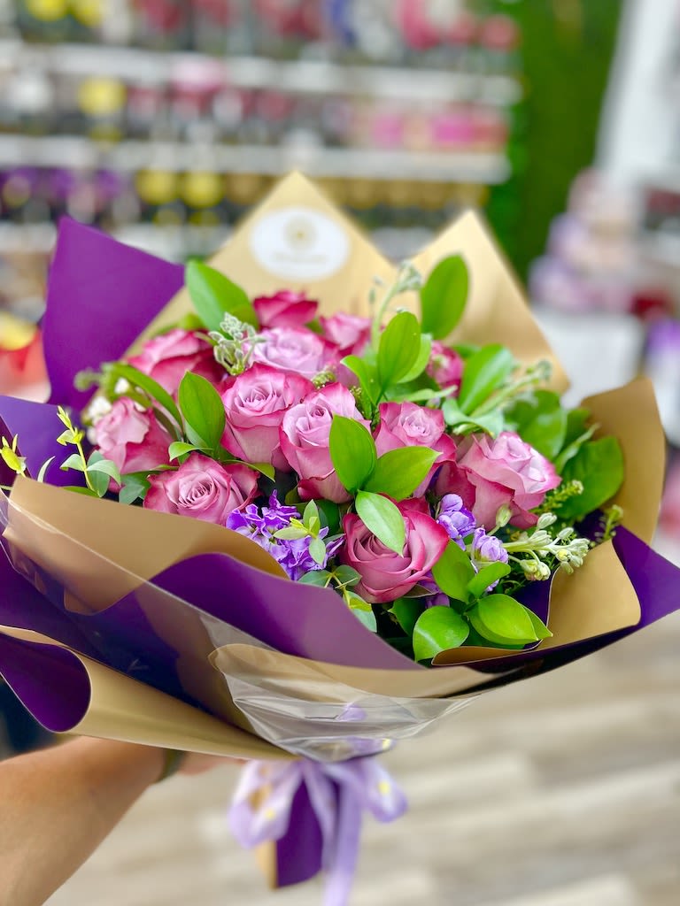 Handheld bouquet of pink roses with purple accents wrapped in tan and purple paper with a ribbon