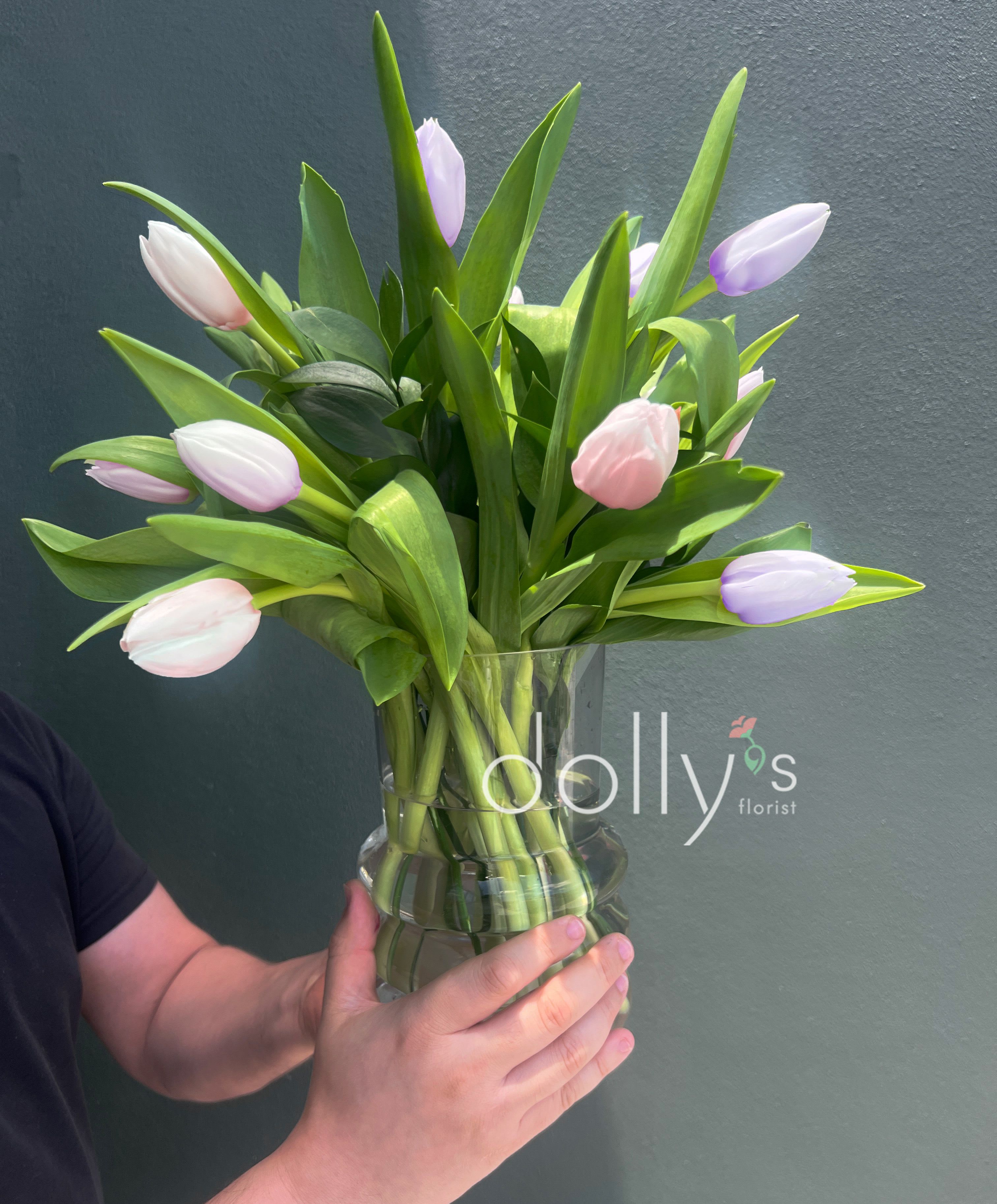 Graceful Gratitude - A fresh cluster of pastel tulips creates a gentle spring moment - perfect as a birthday bouquet, a cheerful get-well gift, or a simple "thinking of you" arrangement. The soft pink, white, and lavender tulip heads sit upright among broad green leaves, so the person you send it to will feel uplifted by its airy, modern charm.  Each arrangement is hand-gathered and presented in a clear glass vase that showcases the clean stems and adds to the arrangement's light, contemporary look. The mix of pale blooms and vivid foliage offers soft color contrast without fuss; tulips are arranged loosely for a natural, garden-fresh silhouette that fits tabletops or entryway cabinets.