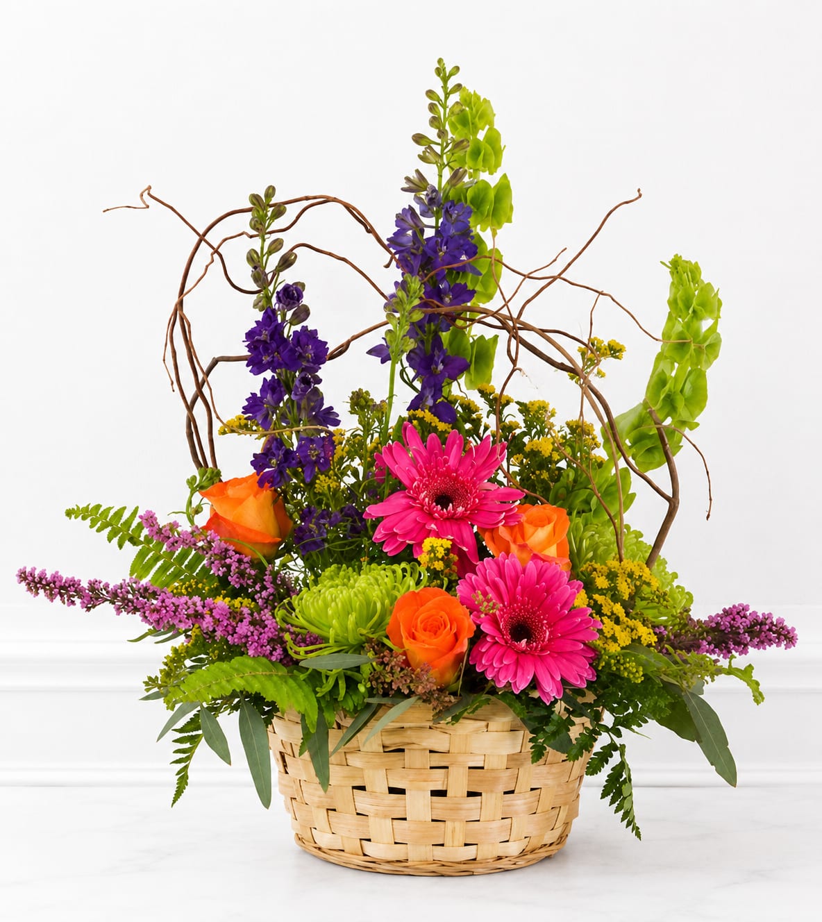 Springtime Basket - A lively burst of color, this Springtime Basket is a cheerful everyday flower arrangement perfect for a birthday, a thank-you, or simply to say "I'm thinking of you." Bright pink gerbera daisies and vivid orange roses take center stage while tall purple blooms and fresh green accents keep the composition energetic and joyful - the kind of gift that makes the recipient smile the moment it arrives.  Arranged in a woven basket, the design mixes saturated color with varied textures: broad gerbera petals, classic rose form, and airy stems topped by curly willow branches that arc above the bouquet for a whimsical, open silhouette. The basket gives it a casual, homey feel and makes the arrangement easy to display on a table, mantel, or entry console.  Ideal for the person who appreciates bold color and playful design, this basket sends a bright, uplifting message without needing a special occasion. No-fuss care keeps it looking fresh: trim stems and change water every few days, and the durable basket presentation makes same-day gifting or delivery simple and charming.