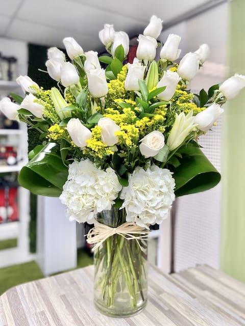 Tall arrangement of white roses, lilies, and hydrangeas with yellow accents in a clear glass vase