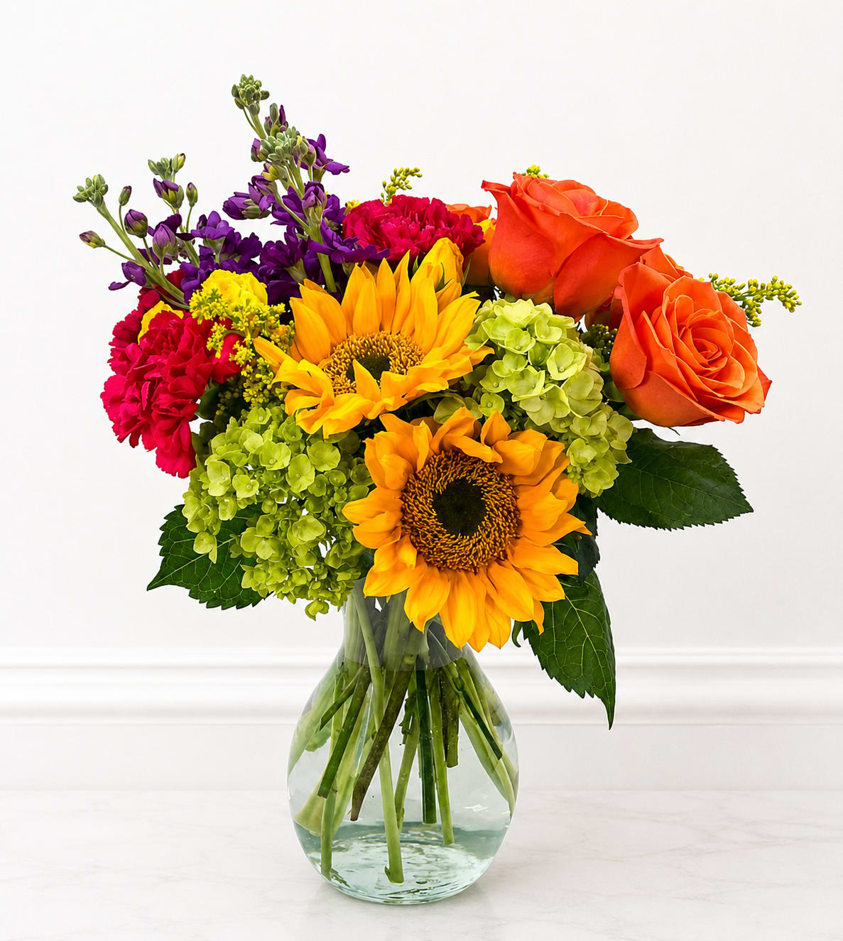 A Smile for You - Vivid orange roses and golden sunflowers take center stage in this cheerful flower arrangement, a lively birthday bouquet or "just because" gift that brightens any room. Lime-green hydrangeas and magenta carnations add lush volume while tall purple stock lifts the composition with height and gentle fragrance-together they create a high-contrast, celebratory look that feels freshly made and joyfully abundant.  Arranged in a clear glass vase, A Smile for You has a full, rounded silhouette that reads bright and generous from every angle. The mix of warm yellows and oranges with cool purples and greens gives the recipient an instant mood lift-perfect for saying thank you, sending get well wishes, or marking congratulations. Sized to sit on a table or bedside dresser, this arrangement arrives ready to display; change the water every few days and trim stems slightly to extend freshness.