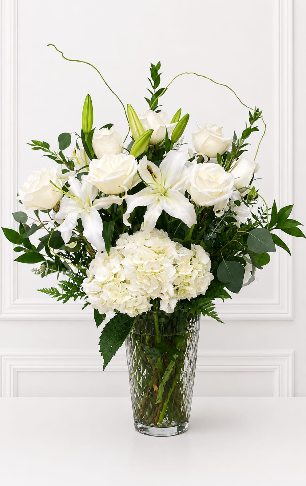Remarkably Perfect - All-white roses, lilies, and a generous cluster of hydrangeas create a serene, graceful centerpiece perfect for an anniversary or a tender "thinking of you" moment. This white floral arrangement for sympathy and funeral or a refined spring celebration reads calm and elegant - the kind of bouquet that comforts the person you're thinking of or marks a milestone with quiet dignity.  Built upright in a tall clear glass vase, the design pairs long lily buds and looping accent stems with dense hydrangea blooms and classic roses for a full yet airy silhouette. Fresh green foliage lifts the white palette, giving subtle contrast and texture; the vase height and vertical lines make it well suited for mantel displays, reception tables, or beside a bedside.   Includes a dozen white roses,  lilies and hydrangeas.