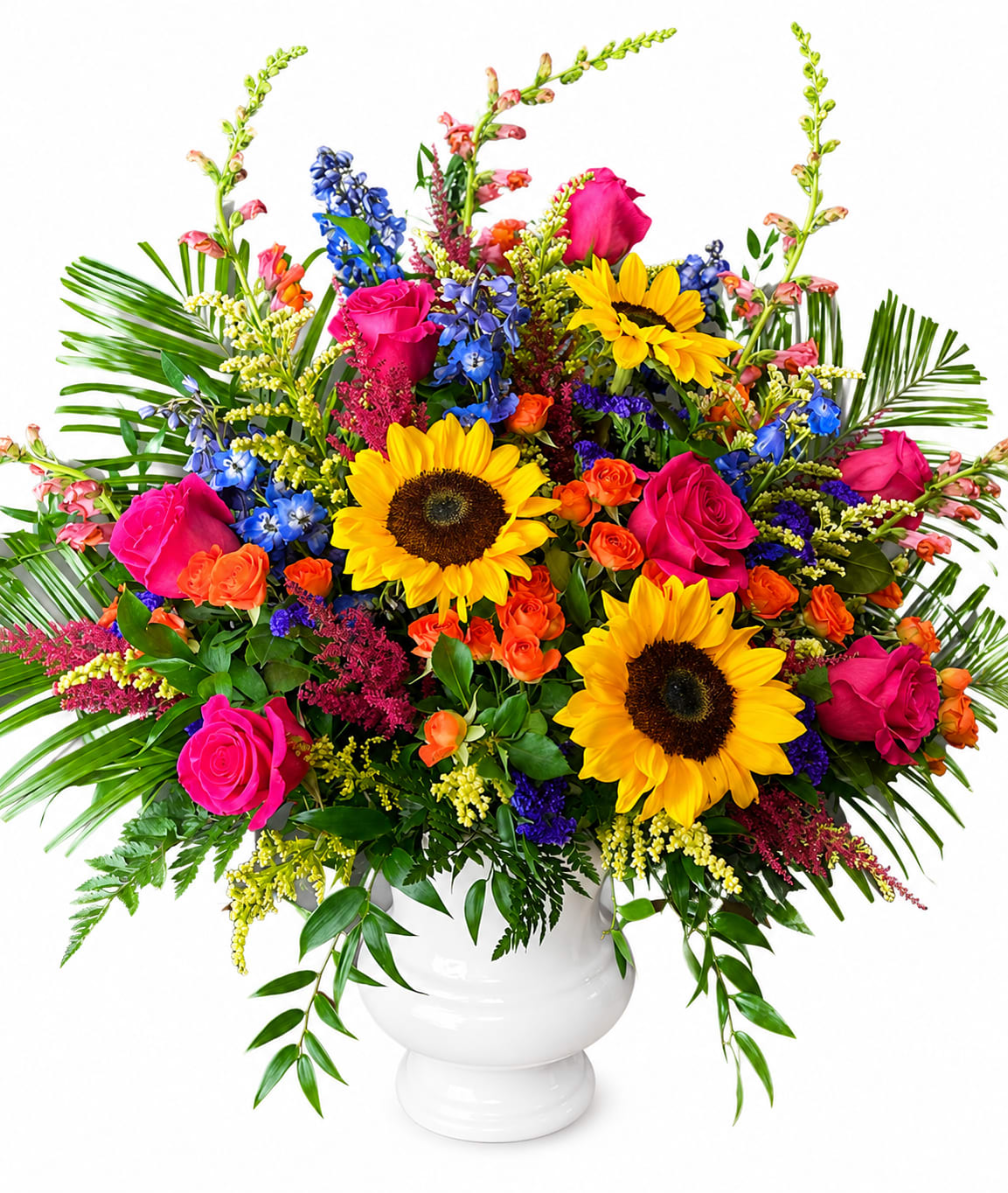 Vivid Blooms Urn - Two bold sunflowers anchor this large, celebratory flower arrangement, the kind of birthday bouquet or congratulations gift that makes the recipient feel instantly seen and uplifted. Vivid pink roses crown the composition, while bright blue spikes and pops of orange lend energetic contrast-together they create a saturated, joyful palette perfect for birthdays, milestones, or a sunny everyday surprise.  Arranged densely in a clean white ceramic vase, the bouquet reads full and expansive with tightly layered blooms across the top and long, arching greenery for dramatic shape. You'll find sunflowers, pink roses, snapdragons and a mix of complementary blooms in yellow, blue, orange and pink tones that play off the vase's simple silhouette.  This arrangement is ideal for celebrating someone who loves bold color and generous floral statements-grandparents, friends, coworkers, or new homeowners. Standard care is to refresh the water every couple of days and trim stems if possible to extend vase life; available in the full, large presentation shown, it arrives ready to brighten the room.