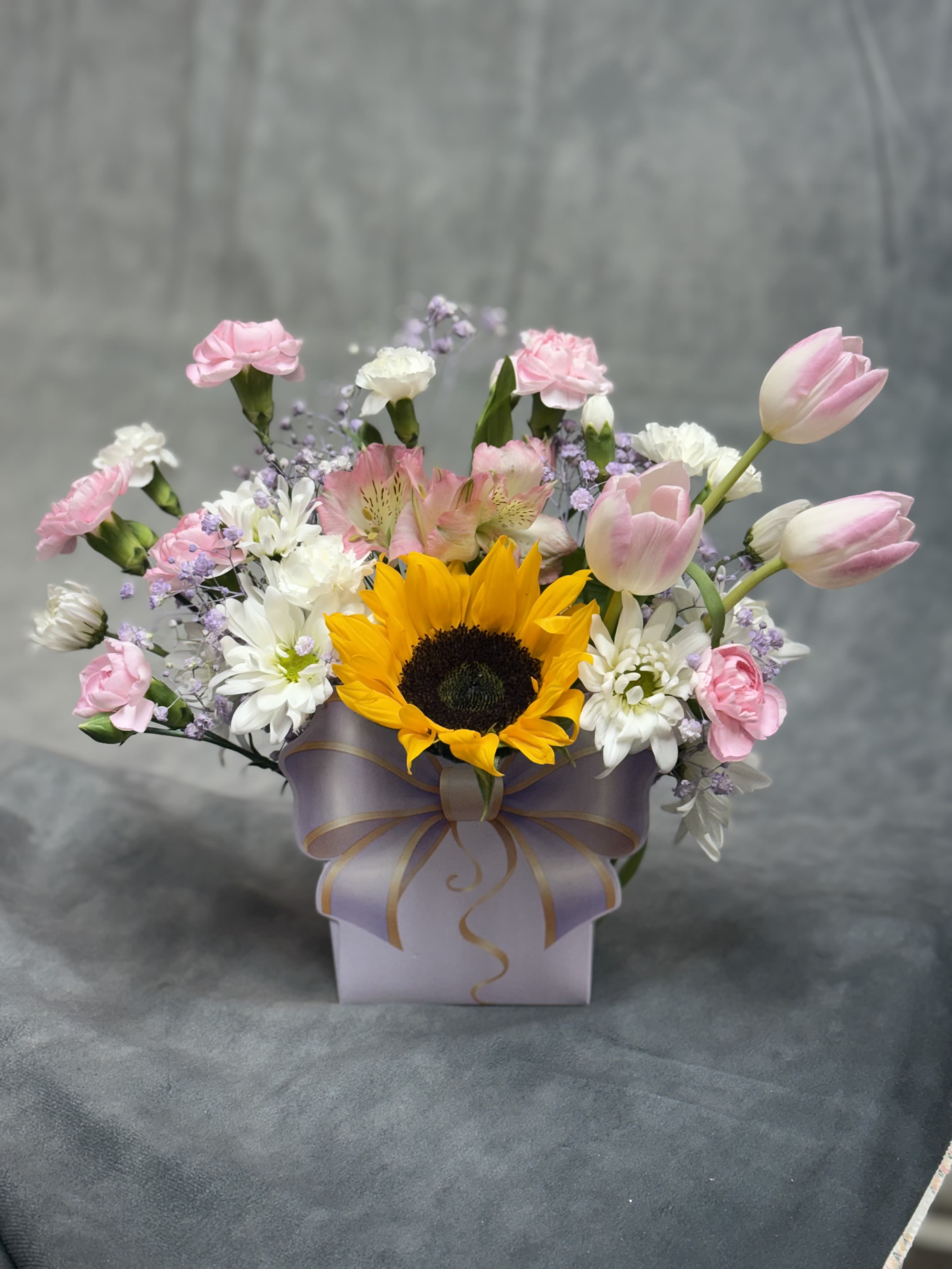 Sunshine Blush Bow Bag  - Bright, cheerful, and full of charm, our Sunshine &amp; Blush Bow Bag features a radiant sunflower surrounded by soft pink tulips, delicate carnations, daisy mums, alstroemeria, and airy lavender accents. Arranged in our signature bow-shaped gift bag, this design brings together sunshine and sweetness to create the perfect gift for birthdays, celebrations, get-well wishes, or just because.  Flowers and accents may vary based on seasonal availability and freshness while maintaining the same overall style, color palette, and value.