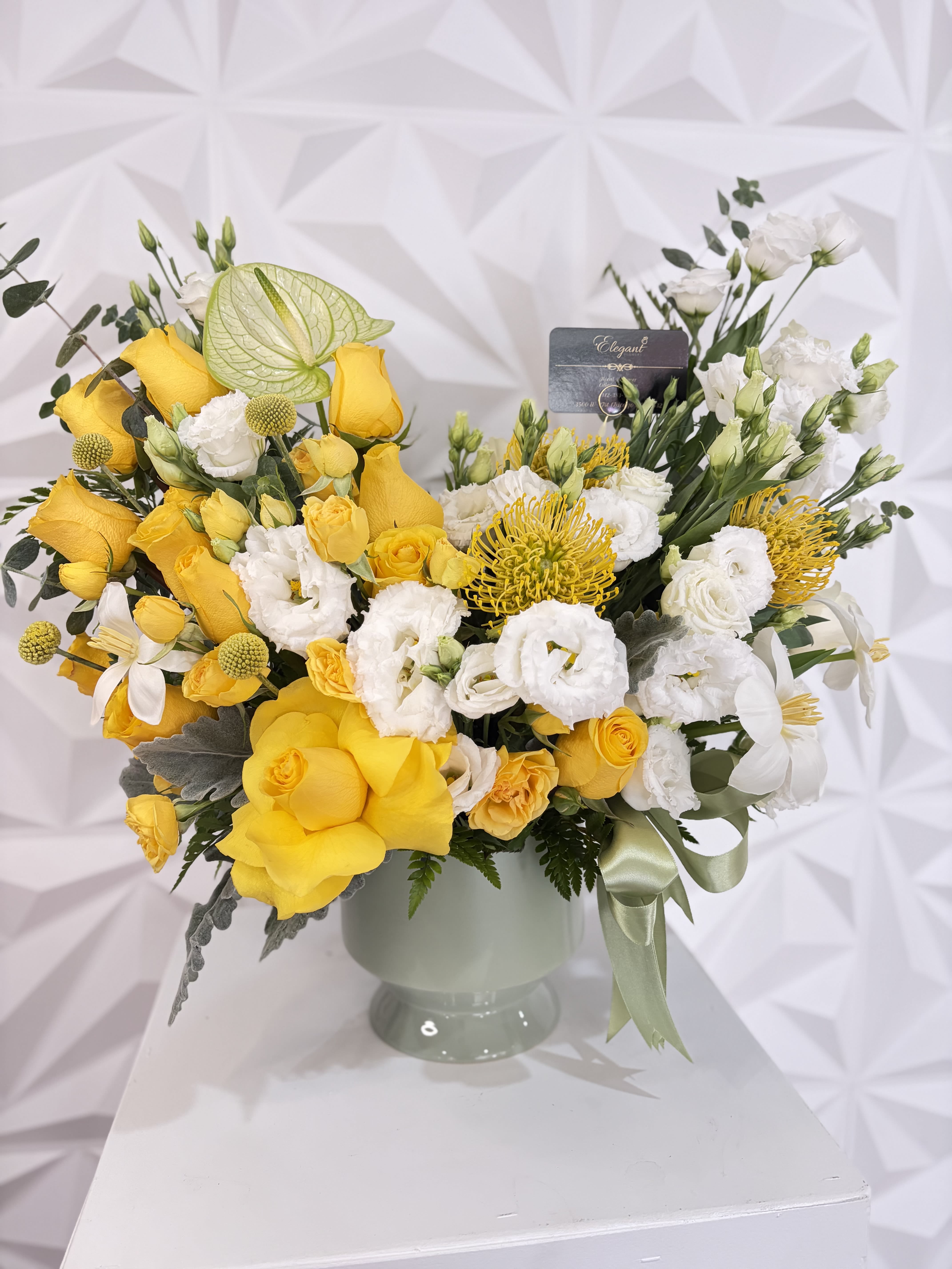 Elegant Flowers collection - This sophisticated floral arrangement is beautifully designed in an elegant ceramic vase, featuring a stunning combination of yellow and white tones that symbolize joy, elegance, and freshness. The design includes beautiful yellow roses, delicate white lisianthus, exotic pincushion proteas, cheerful craspedia, elegant green anthuriums, and a distinguished yellow tulip, complemented by lush greenery that adds texture and movement. 