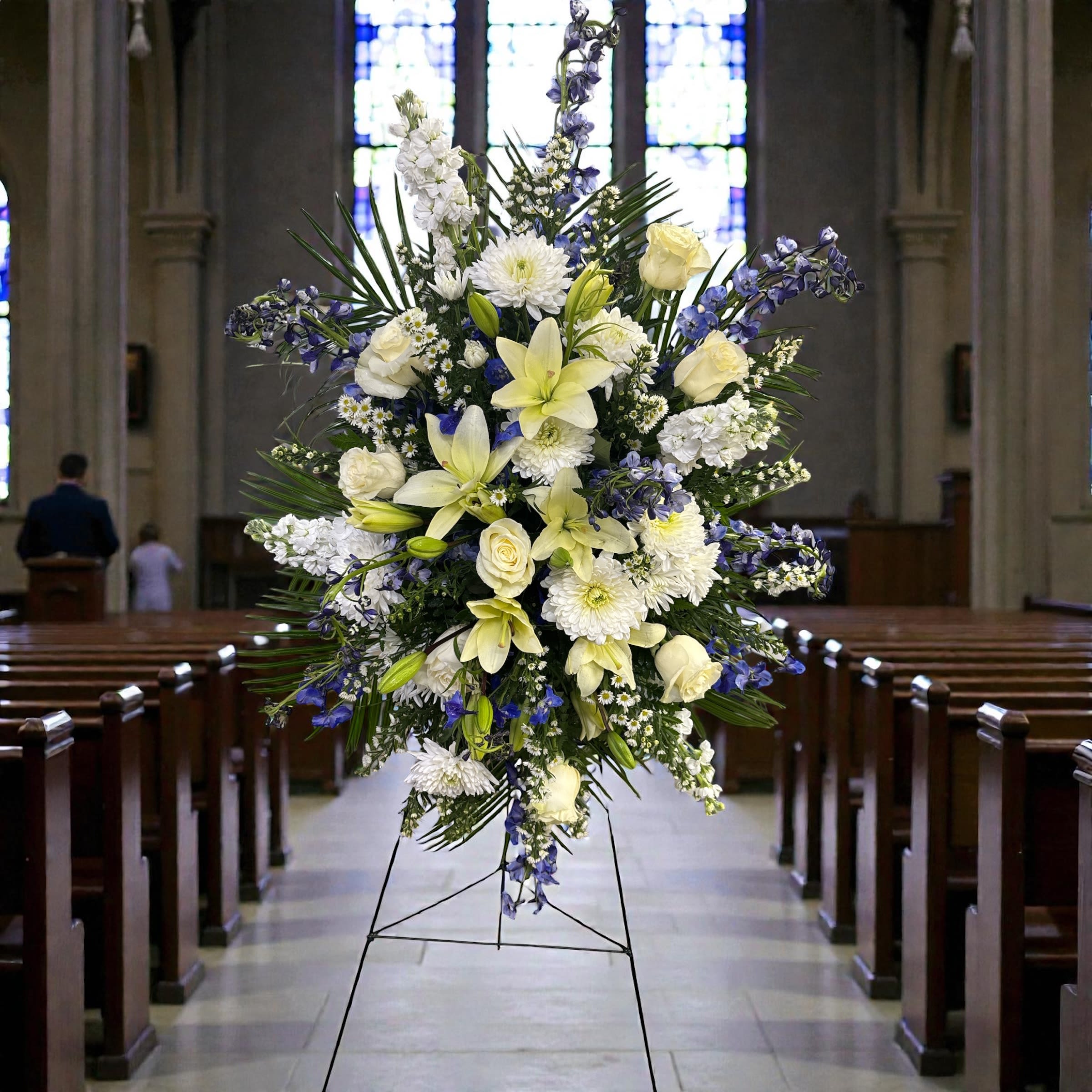 Faith and Remembrance  - This white and blue standing spray is designed to bring a sense of peace, comfort, and quiet presence to the service. A classic combination of white lilies and roses is accented with soft blue tones, creating a design that feels calm, balanced, and respectful without being overly traditional.  The vertical shape gives it natural height and visibility, allowing it to stand with presence while still maintaining a gentle, composed feel appropriate for the setting.  A meaningful choice for funeral services, memorial services, or celebration-of-life gatherings, offering a tribute that honors a loved one with dignity and timeless beauty.  Each arrangement is created with the freshest seasonal stems available. Subtle variations may occur while preserving the overall style, color palette, and design.  Available for standing spray delivery through Aspen Tower Florist, serving Shelbyville and Greensburg along with Rushville, Batesville, Westport, Oldenburg, St. Paul, Waldron, and Milroy.