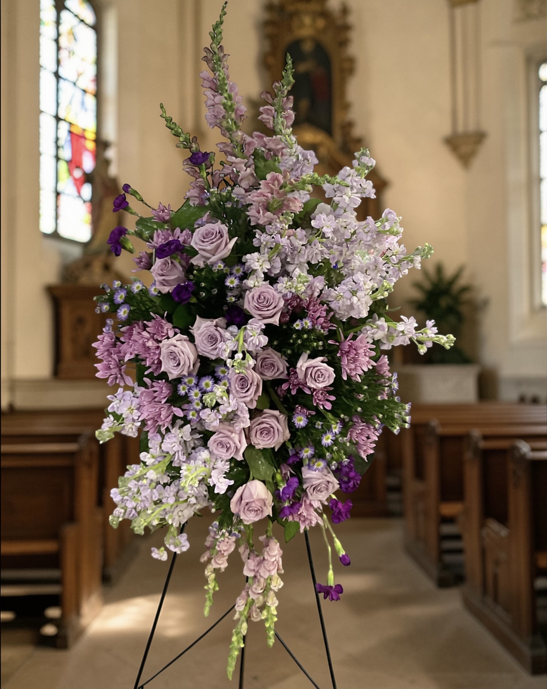Lavender Embrace - Soft, soothing, and beautifully expressive, Lavender Embrace is a heartfelt standing spray designed to surround your loved one’s service with calm, comforting color.  Lavender roses, fragrant stock, snapdragons, mums, and accent blooms are layered in rich tones of lilac, plum, and soft purple, creating a lush, full presentation that feels both uplifting and serene.  The vertical design offers natural height and presence, allowing it to stand gracefully at the service while maintaining a gentle, composed feel appropriate for the setting.  This piece is ideal for families who want something refined, elegant, and deeply personal—an expression of love that feels peaceful without being understated.  Each arrangement is created with the freshest stems available. Subtle variations may occur while preserving the overall color palette and design.  Available for standing spray delivery through Aspen Tower Florist, serving Greensburg and Shelbyville along with Rushville, Hope, Batesville, Westport, Oldenburg, St. Paul, Waldron, and Milroy.