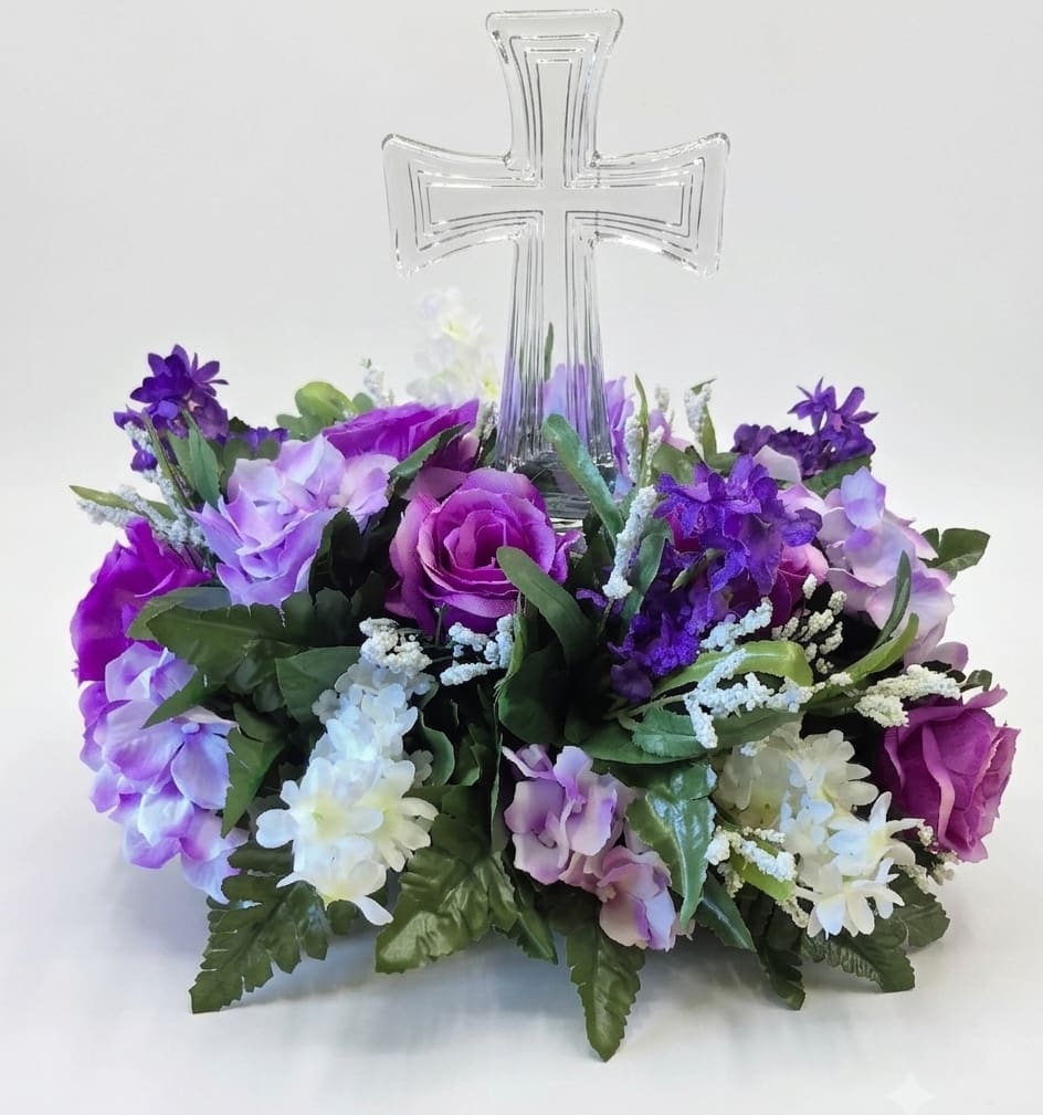 Crystal Cross with Silk Shades of Purple Flowers  - A clear crystal cross surrounded by a halo of silk blooms creates a calm, reverent sympathy arrangement that honors memory with quiet elegance. This flower arrangement for memorials and funerals features shades of purple, lavender, and white to convey comfort and devotion, the kind of tribute that helps the person you're remembering feel respectfully celebrated.  The composition is densely packed with silk roses, hydrangea clusters, and small white accents set against soft green foliage for a layered, full appearance. The crystal clear cross stands at the center and remains visible through the blooms, giving a devotional focal point while the mixed textures of petals and clusters add gentle visual depth. Colors are balanced so the purples read soothing rather than bright, suited to solemn occasions.  Sized approximately 12" tall and 12" across, this keepsake arrangement holds its shape without wilting and requires no water or maintenance, making it ideal for display at a memorial service, on a gravesite, or as a long-lasting remembrance in the home. Choose this piece to send condolences and offer a thoughtful, lasting expression of sympathy. Also would be a great gift for Get Well, Birthday or Just Because. 