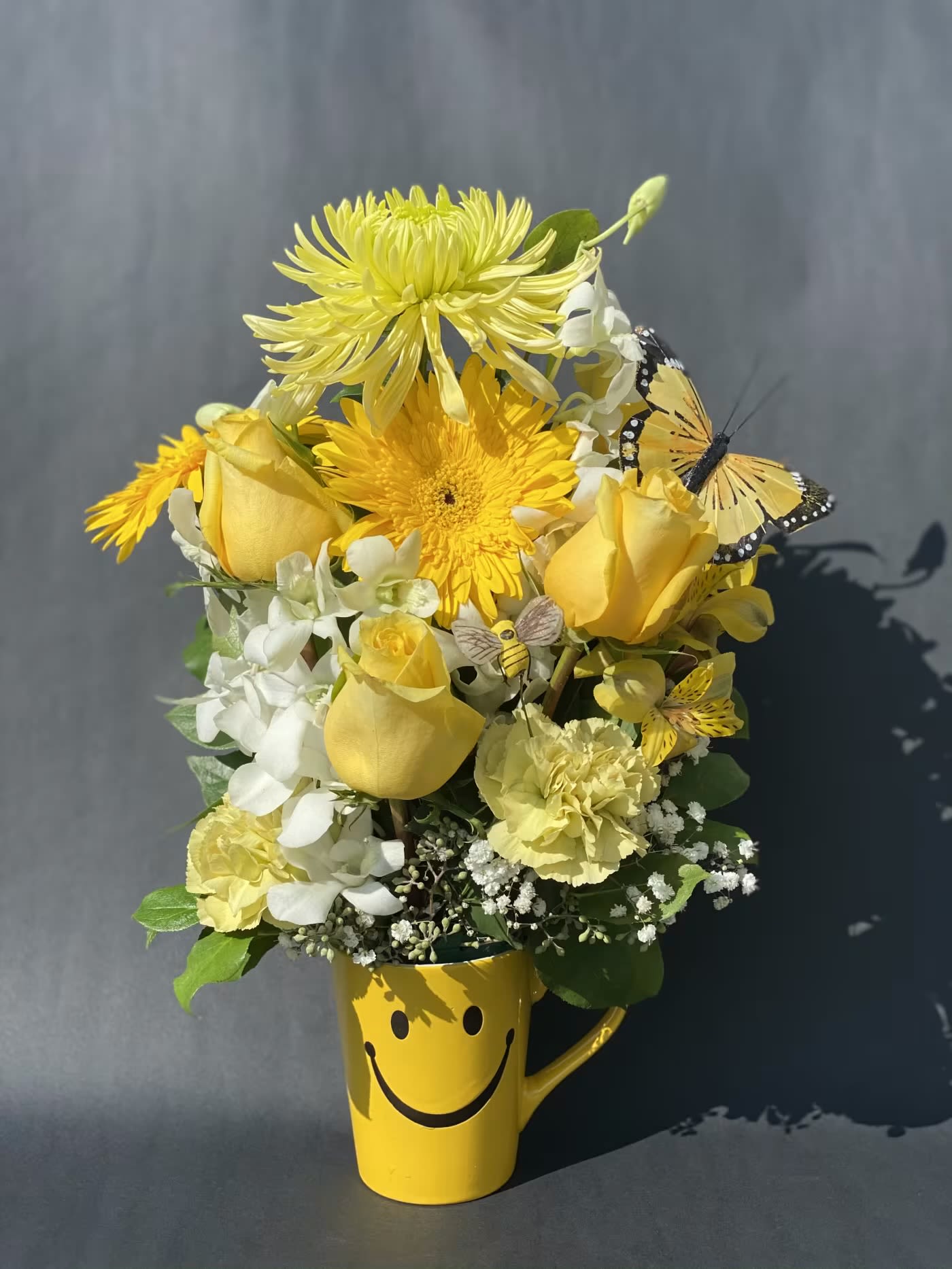 Yellow flower arrangement in a smiley-face mug with butterfly accents