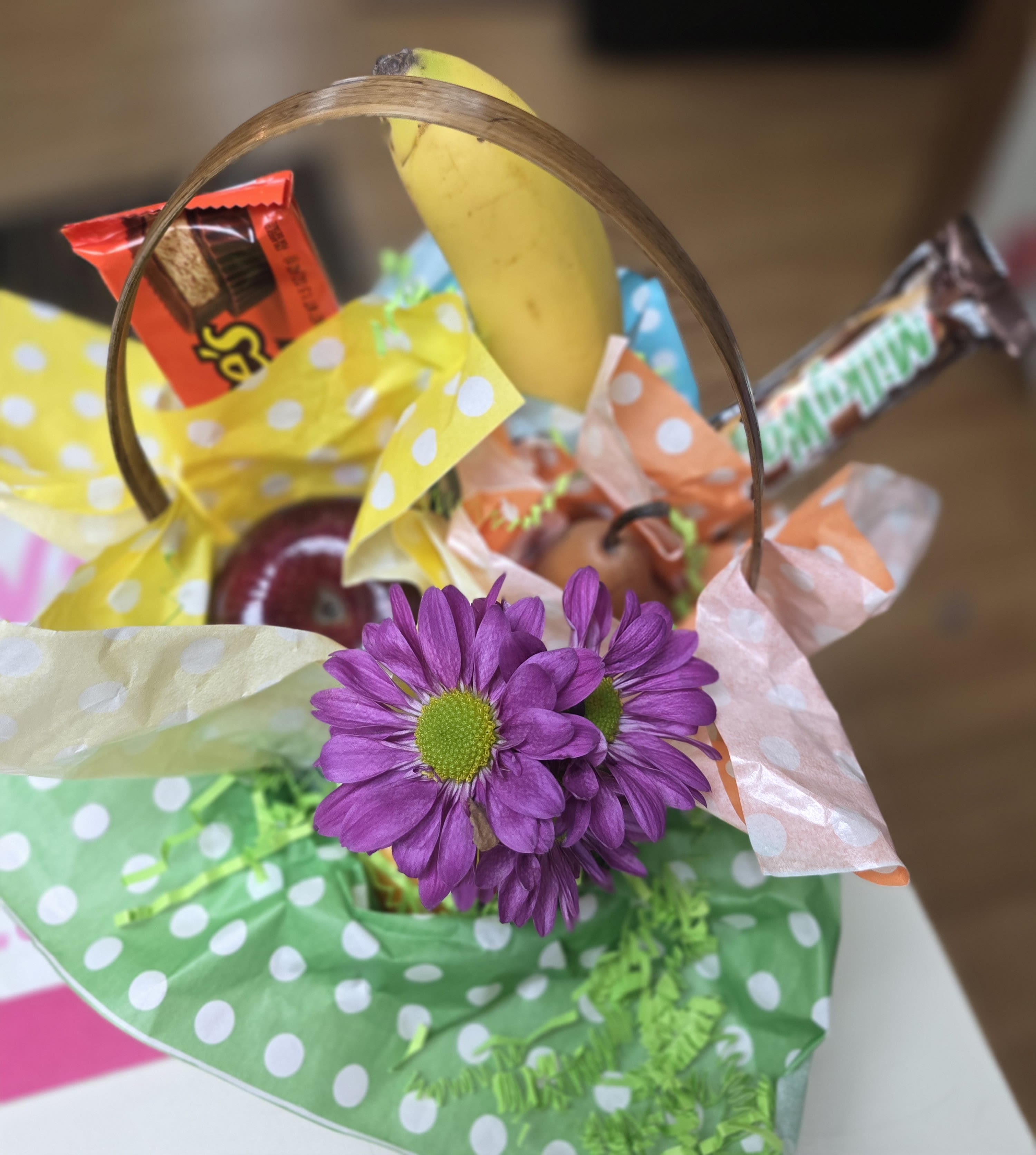 FBDD Snack Basket - A playful snack basket that feels like a cheerful surprise-this flower arrangement-style gift mixes purple daisy blooms with fresh fruit and wrapped candy in a small handled basket, perfect as a birthday gift, thank-you surprise, or just-because treat. The bright polka-dot tissue paper in yellow, green, peach, and orange gives the presentation a sunny, casual vibe so the recipient will feel celebrated the moment they see it.  Inside the basket you'll find several purple daisies tucked among a selection of fruit (banana and apple) and assorted candy bars, all arranged on colorful polka-dot tissue for a fun, gift-ready look. The arrangement reads more like a mixed gift basket than a traditional bouquet, making it ideal for someone who enjoys sweet treats and a fresh floral accent.  Give this to a friend, co-worker, or family member who prefers snacks over formal flowers-the combination of fruit, candy, and bright blooms sends a lighthearted, thoughtful message. The compact handled basket is ready to present; no extra wrapping needed.