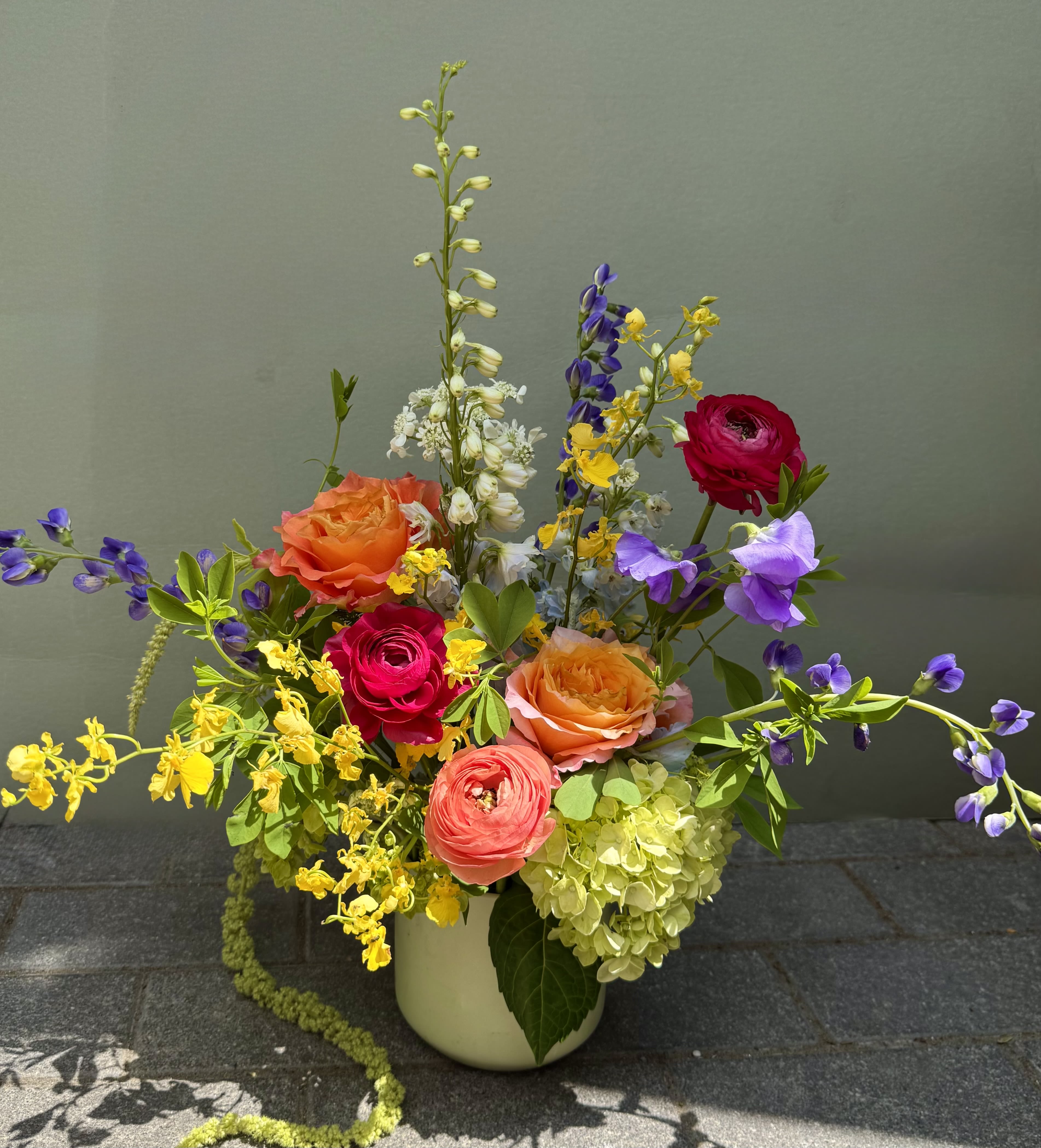 California  - A lively garden-style mixed bouquet that bursts with warm pinks, corals and bright oranges balanced by cool purples and sunny yellows - perfect as a birthday bouquet, congratulations arrangement, or an everyday gift to make someone feel celebrated. The layered composition places bright roses and ranunculus front and center so the recipient will notice their full, velvety blooms first, while taller, airy stems and delphinium add movement and a joyful silhouette that reads fresh and unstructured.  This hand-tied look is arranged into a clean white ceramic vase and anchored by a pale green hydrangea cluster for texture and volume. You'll also find delphinium spires and other mixed accent blooms that give height, contrast, and a slightly whimsical, romantic touch. Colors include mixed bright pink, coral, purple, yellow and orange tones for a saturated, springlike palette.  Ideal for friends, mothers, coworkers, or partners who love color and a garden-fresh feel - the kind of bouquet that makes someone feel truly seen on their special day or simply because you're thinking of them. Vase included; arrange out of direct heat and change the water every 2-3 days to keep blooms looking their best.