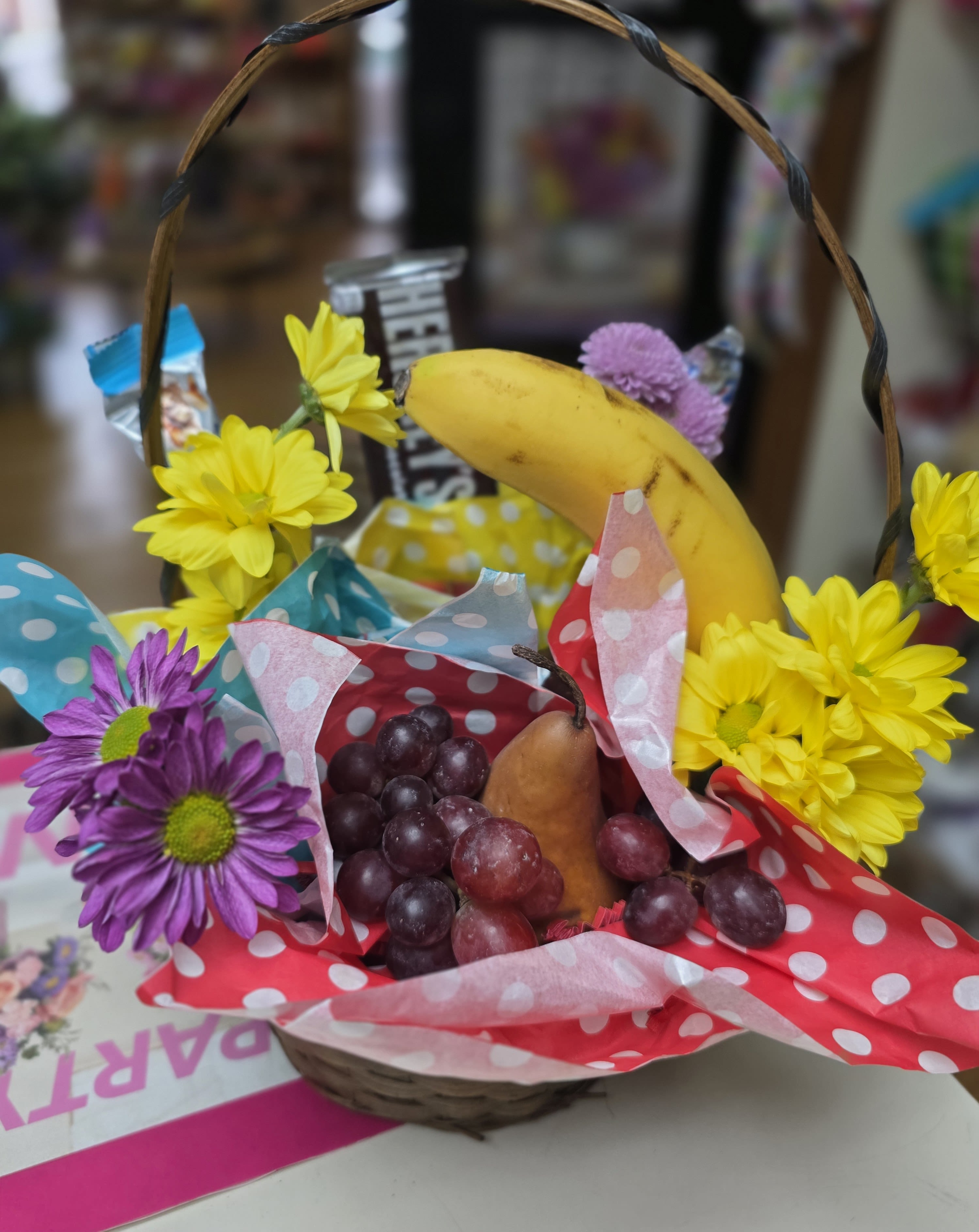 Fruit &amp; Chocolate Gift Basket - Bright yellow and purple daisies peek out from a woven basket brimmed with fresh fruit, offering a sunny, cheerful gift for birthdays, thank-yous, or a just-because pick-me-up. This fruit and chocolate gift basket reads as playful and abundant-the colorful blooms and polka-dot tissue paper give it a handmade, festive feel that will make the person you're thinking of smile the moment it arrives.  Inside the basket you'll find a ripe banana, pear, and clusters of red grapes nestled alongside daisies in yellow, purple, pink, and red tones, with premium candy bars tucked into the arrangement for a sweet finish. The woven basket handle gives it a casual, giftable look and the tissue paper adds a pop of pattern-perfect for a cheerful delivery that feels thoughtful without being formal. Ideal for coworkers, friends, or family who appreciate a bright, edible surprise; sizes and exact fruit selection may vary slightly with seasonality.