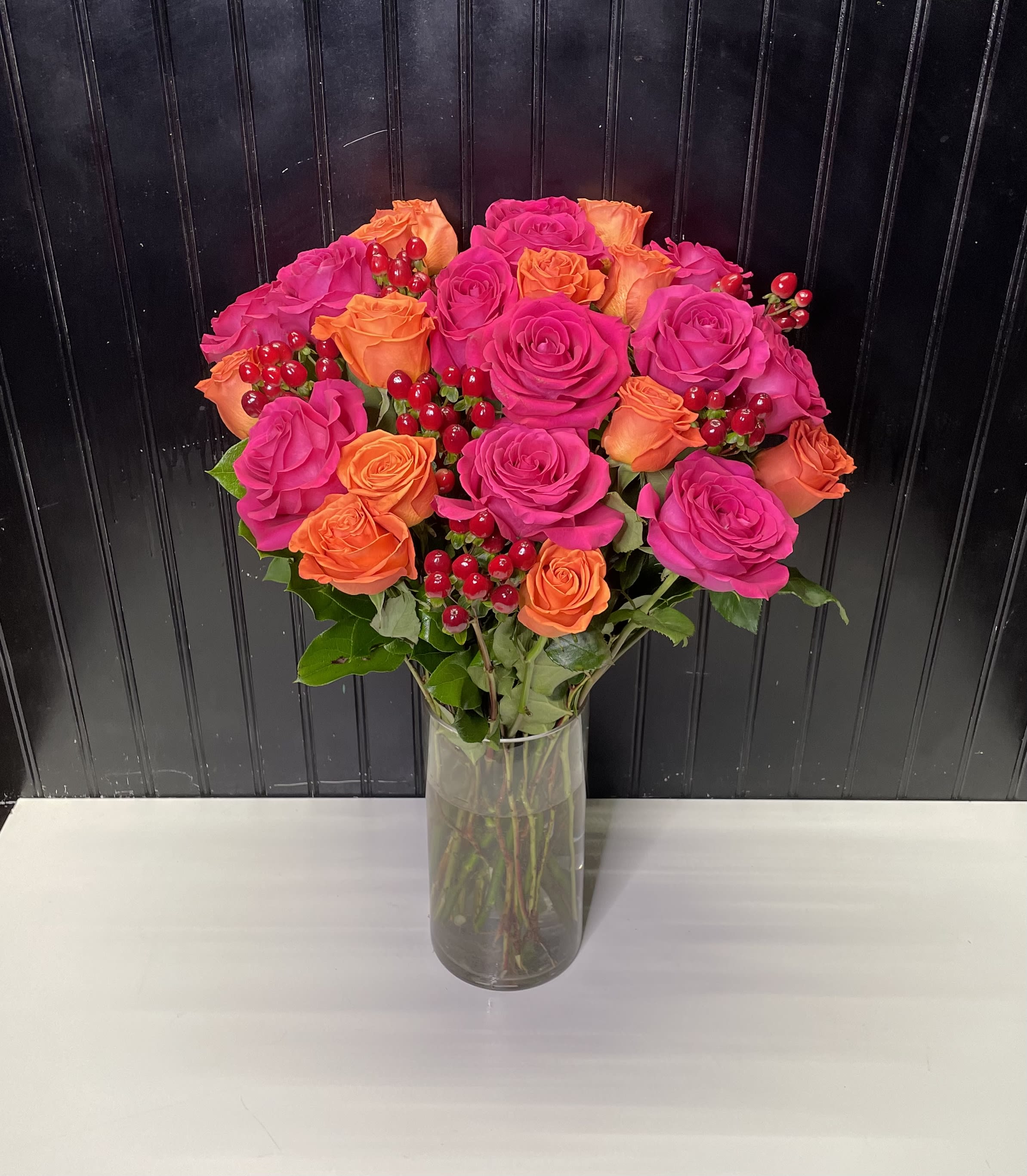 Tall glass vase of hot pink and orange roses with red berries in a rounded bouquet