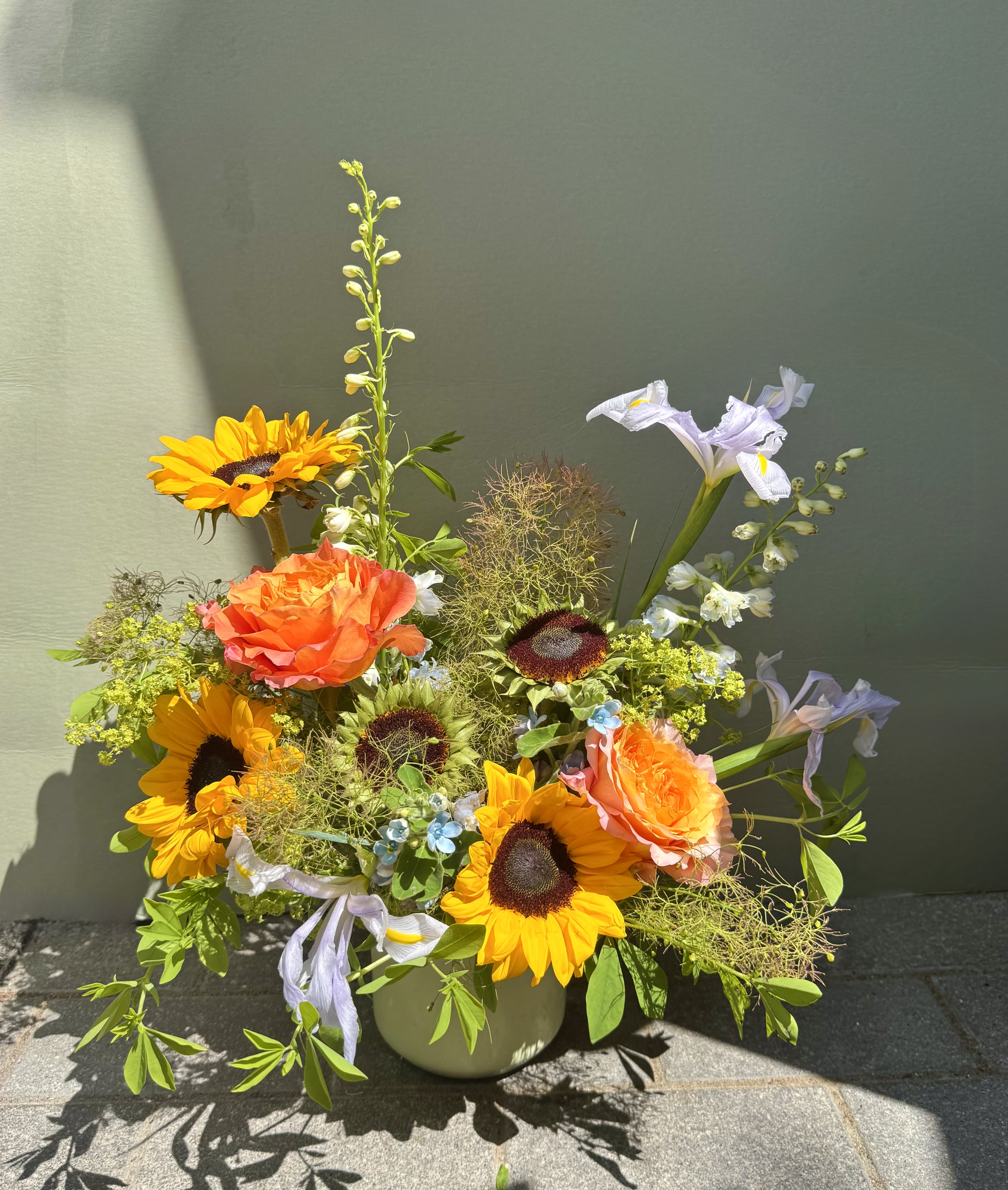 Galapagos - Bright sunflowers take center stage in this Galapagos flower arrangement, a warm, cheerful bouquet perfect as a birthday, thank-you, or spring celebration gift. The mix reads sunny and playful-yellow sunflowers and orange roses bring bold, joyful color while pale lavender irises and small blue accents add a soft, whimsical contrast that feels hand-picked from a cottage garden. The overall effect is lively and heartfelt, the kind of arrangement that makes the person you're thinking of feel genuinely celebrated.  This garden-style composition is arranged in a simple ceramic vase, with stems left airy and slightly untamed so blooms extend in different directions for a natural, relaxed silhouette. Textural filler and wispy greens add movement and depth while highlighting the striking shapes of the sunflowers, roses, and irises. Sized to sit on a table or desk, it's an inviting centerpiece that smells subtly floral and looks fresh for days-an effortless, joyful way to mark a happy occasion.