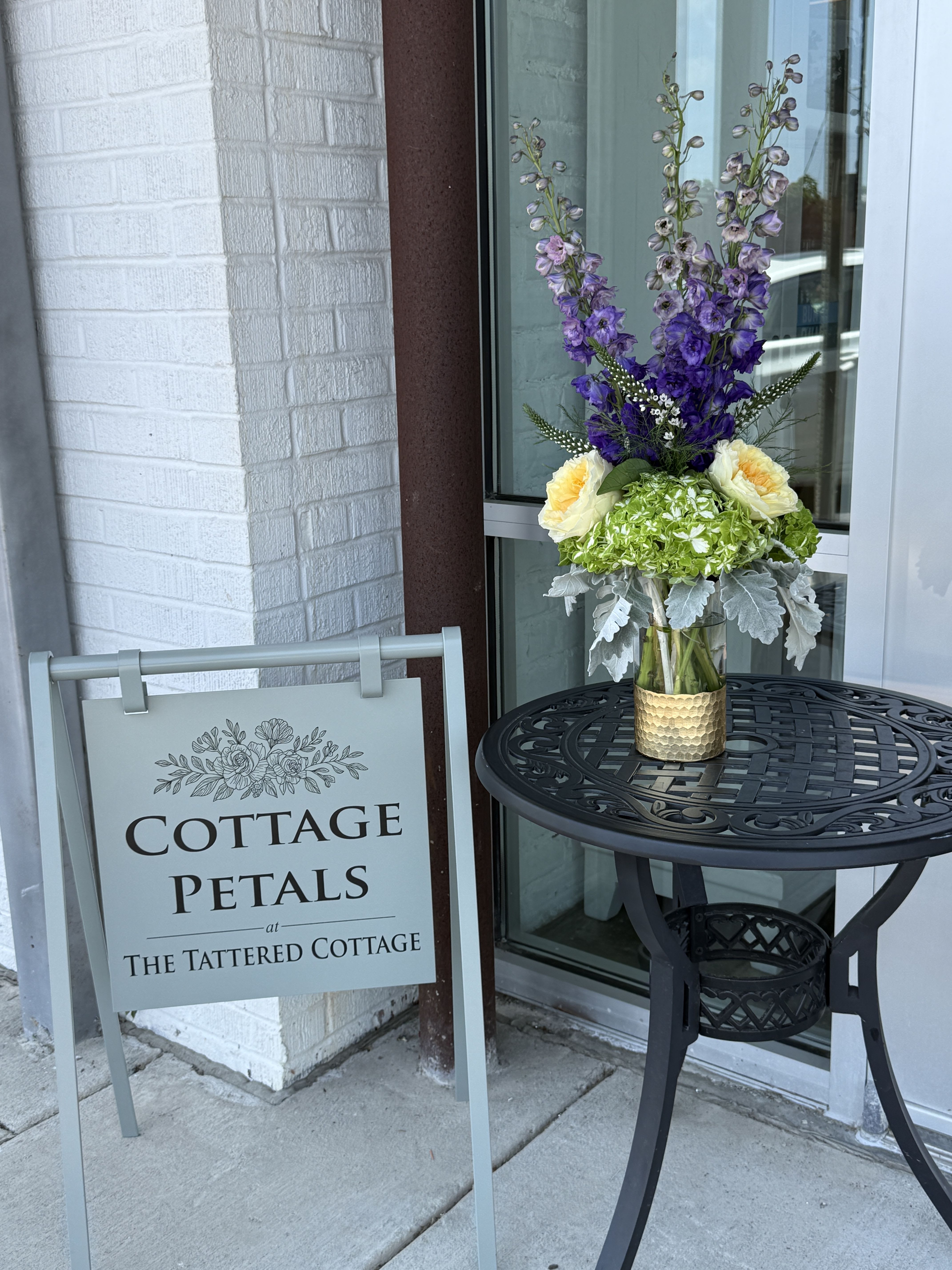 Lavender Meadow Glow - A striking blend of rich purples, soft creams, and fresh greens, Lavender Meadow Glow brings the beauty of a blooming countryside garden indoors. Featuring tall lavender delphinium, lush green hydrangea, creamy garden roses, delicate veronica, and soft dusty miller foliage, this arrangement offers graceful height, texture, and movement. Designed in a modern gold-accented vase, it makes a beautiful gift for birthdays, celebrations, sympathy, or simply brightening someone's day with natural elegance.  Flower varieties and colors may vary based on seasonal availability while maintaining the overall style, color palette, and value of the arrangement.