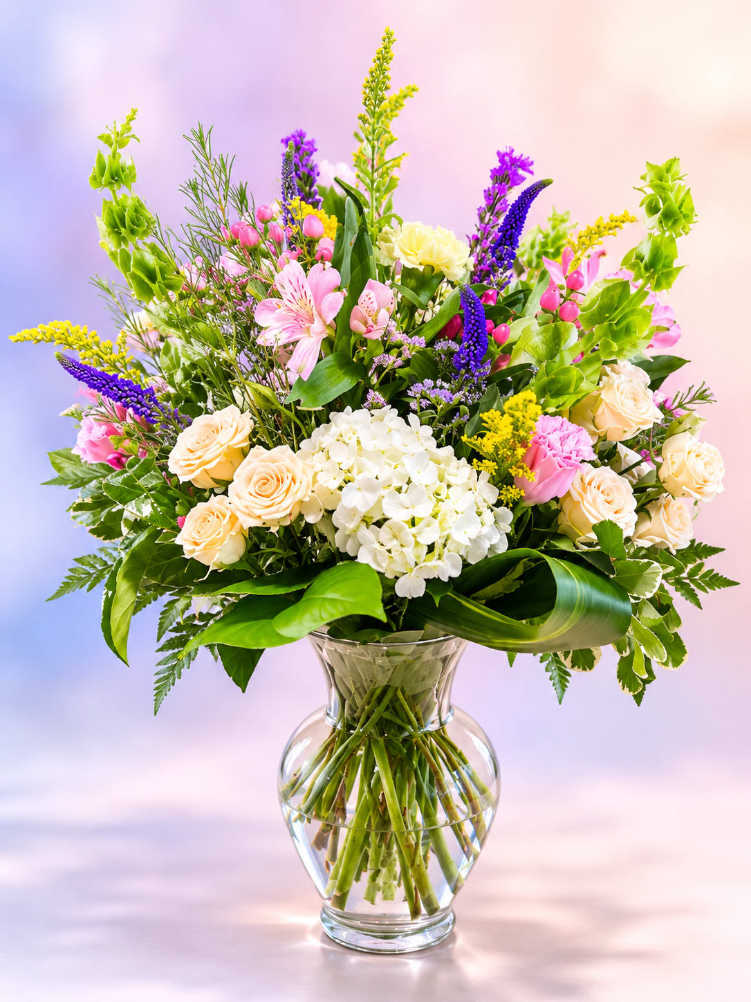 Petals &amp; Promises  - Soft, romantic, and overflowing with garden charm, Petals &amp; Promises is a graceful arrangement designed to celebrate life's sweetest moments. Featuring creamy ivory roses, delicate pink alstroemeria, fluffy white hydrangea, vibrant purple veronica, cheerful golden solidago, and playful pink hypericum berries, this lush bouquet is artfully gathered with fresh greenery in a clear glass vase. With its airy, meadow-inspired style and timeless beauty, Petals &amp; Promises is a heartfelt expression perfect for birthdays, anniversaries, celebrations, or simply letting someone know they're loved.