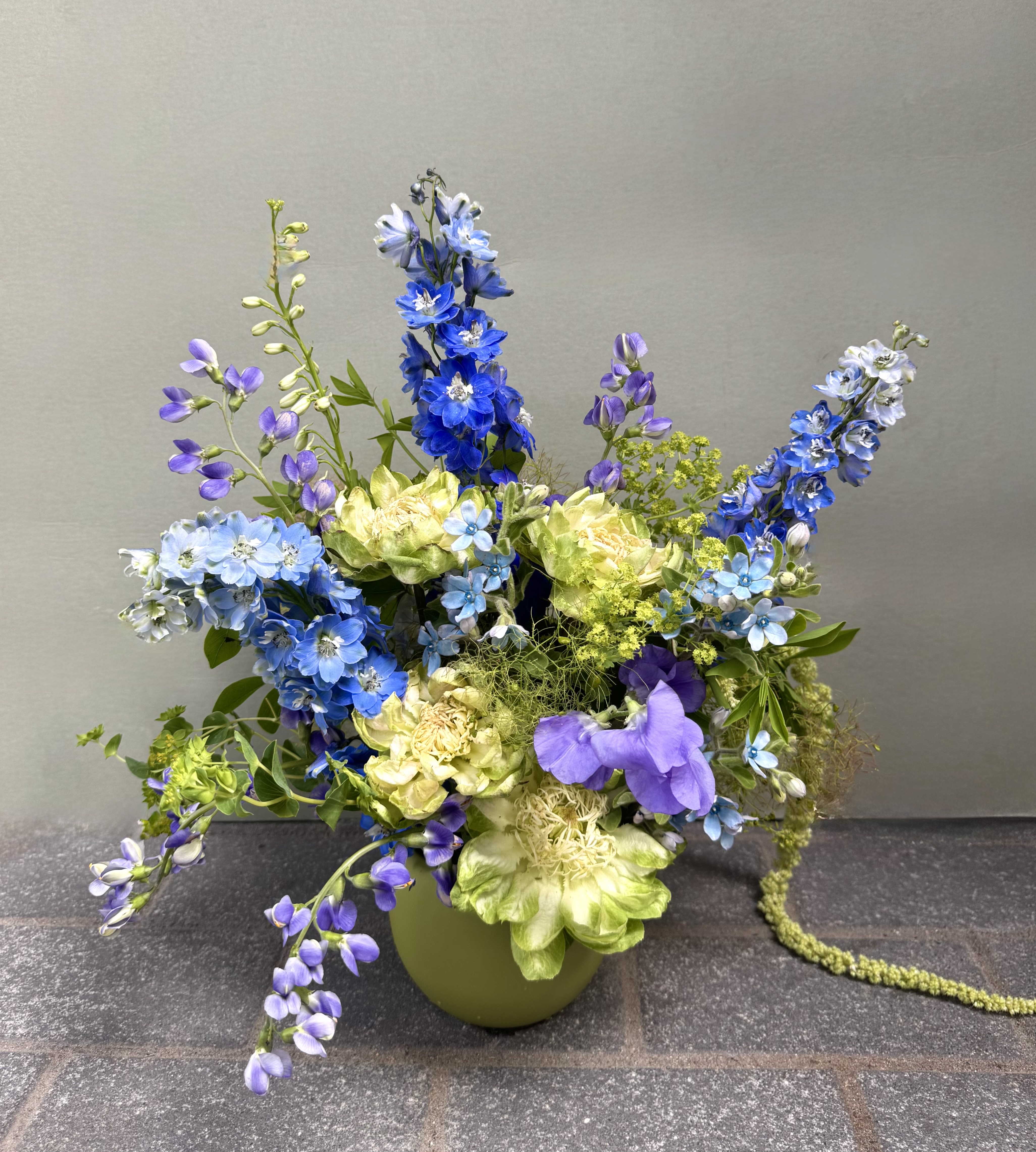 Greece  - Tall spikes of blue and lavender delphinium set a dramatic, airy tone in this garden-style flower arrangement for birthdays, spring gatherings, or thoughtful celebrations. The cool blue and purple palette - echoed in rounded blue hydrangea clusters and small blue accent blooms - feels fresh and elegant, while touches of yellow-green filler bring a hint of sunshine that keeps the design lively rather than formal. The overall look reads whimsical and uplifting, the kind of bouquet that will make the person you're thinking of feel genuinely seen.  This mixed bouquet pairs delphinium with pale green roses and other soft green blooms, layered with airy greenery for movement and texture. Arranged asymmetrically in a low green ceramic vase, it features tall vertical elements balanced by rounded clusters to create both height and fullness without appearing stiff. The cool tones of blue, purple, green, and white mingle with subtle yellow-green accents for contrast and depth.  Perfect for spring or summer occasions - from birthdays and thank-you gestures to just-because celebrations - this arrangement arrives ready to display in its ceramic vase. Its loose, garden-style composition suits recipients who appreciate a natural, slightly whimsical aesthetic; water the stems as needed and keep away from direct heat to prolong freshness.