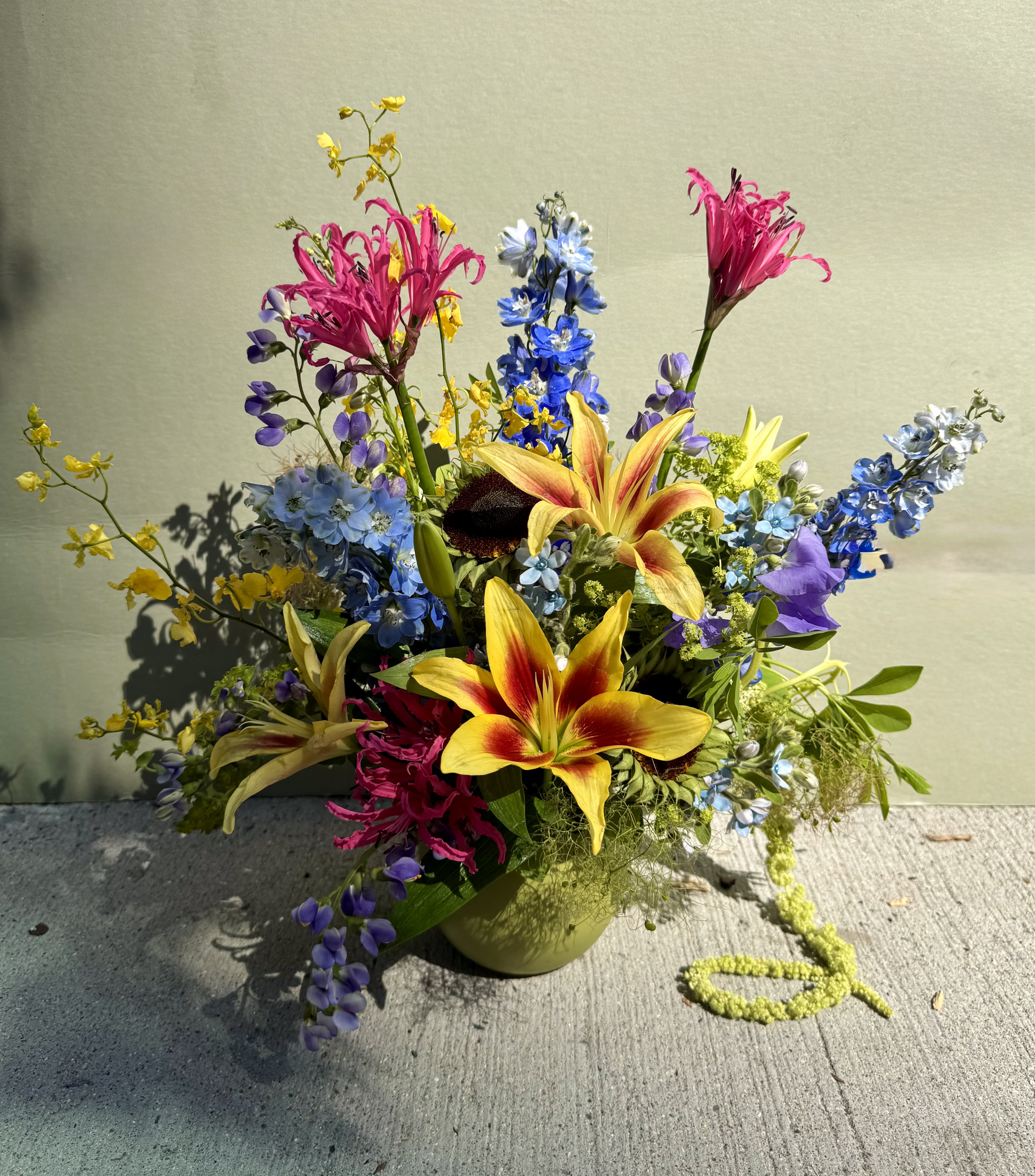 Tropicana  (Designer’s Choice)  - Vivid yellow-and-red lilies take center stage in this mixed-color flower arrangement, creating a bold, celebratory focal point perfect for a birthday bouquet, congratulations gift, or cheerful get-well surprise. Bright blue delphinium spikes and pink blooms lift the composition upward while small purple accents add depth, so the person you're thinking of will feel seen and uplifted the moment they look at it.  The design reads loose and airy, with stems reaching outward for a lively, garden-like silhouette that feels freshly gathered. Lilies, delphinium, orchids, and complementary accent blooms are arranged in a small ceramic vase that adds a clean, modern touch and keeps the display desk-friendly. Saturated yellows, pinks, blues, and purples work together for a dramatic yet approachable palette that suits many celebratory moments.  Ideal for sending to brighten a home, office, or bedside, this arrangement is the kind of gift that says "I'm cheering for you" without needing words. Because it arrives in a ready-to-display ceramic vase, it's easy to place and enjoy right away-simply trim stems and refresh water as needed to help blooms last longer.