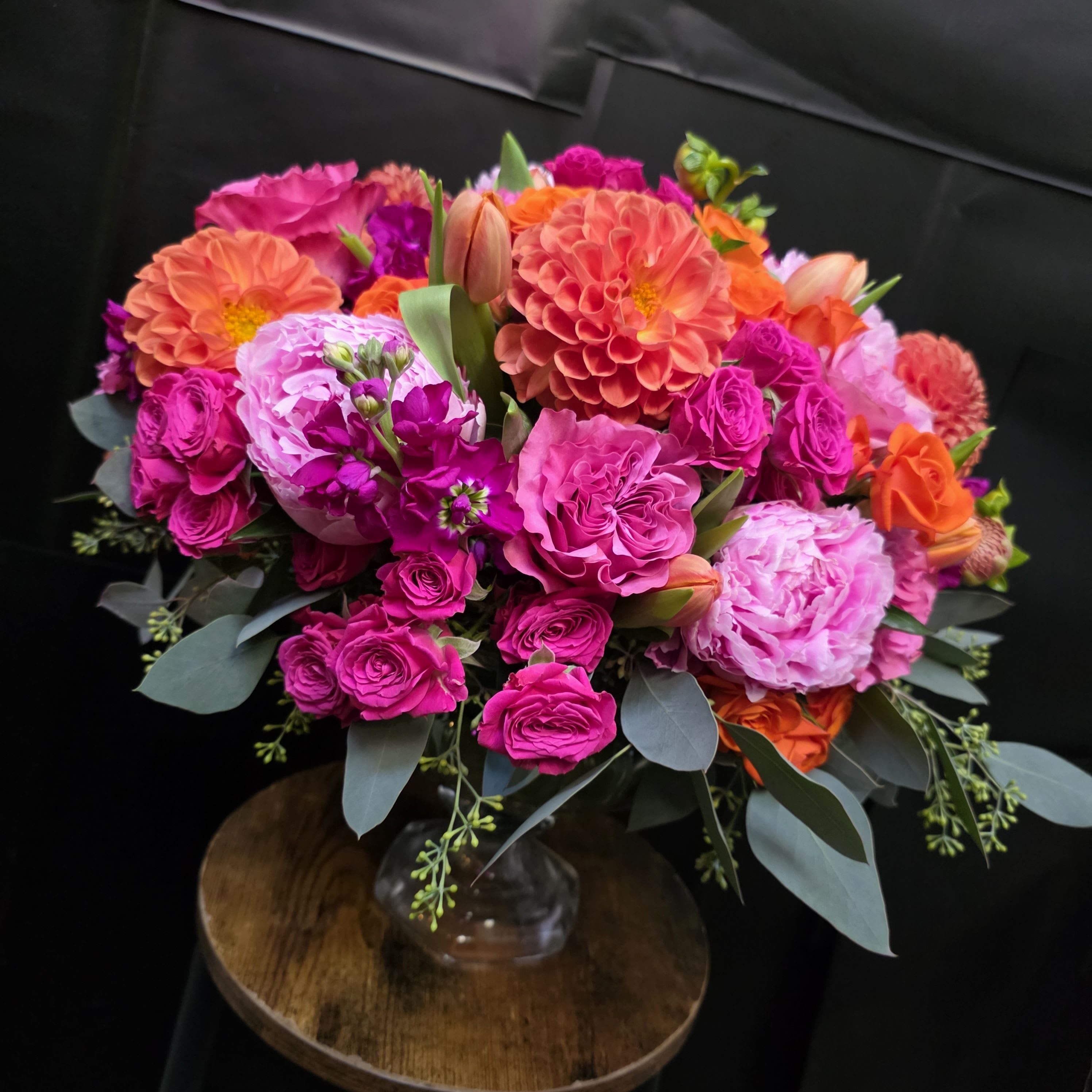 Mia's Pink Passion - A lush, rounded celebration of color, this flower arrangement is a joyful everyday bouquet perfect for birthdays, congratulations, or a thoughtful "just because" surprise. Mia's Pink Passion places large dahlia-like blooms front and center alongside layered roses and tulip buds so the person you're thinking of will see vibrant pinks, coral-oranges, lavenders, and touches of white the moment the vase is set down-an instant mood lifter that reads as both modern and exuberant.  The design is densely packed for a full, textured look: sizeable dahlias provide bold focal points, medium roses add softness and depth, tulip buds and smaller accent blooms create pleasing shape variation, and soft gray-green foliage frames the palette. Arranged in a clear glass vase, the mix of pink, orange, purple, and mixed-color flowers reads bright and celebratory while maintaining a polished, contemporary finish.  Ideal for cheering someone up, saying thanks, or marking a happy milestone, this bouquet feels personal and festive so she'll feel genuinely celebrated. The vase arrangement arrives ready to display; simple care-fresh water and a cool spot away from direct sun-keeps blooms looking their best for days.