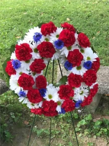 USA Wreath Spray - Celebrate Independence Day with timeless elegance through the USA Wreath Spray, a design that honors the American spirit in vibrant color and texture. Each wreath includes red carnations, blue delphinium, and white mums in a balanced, circular composition that symbolizes freedom, unity, and celebration.