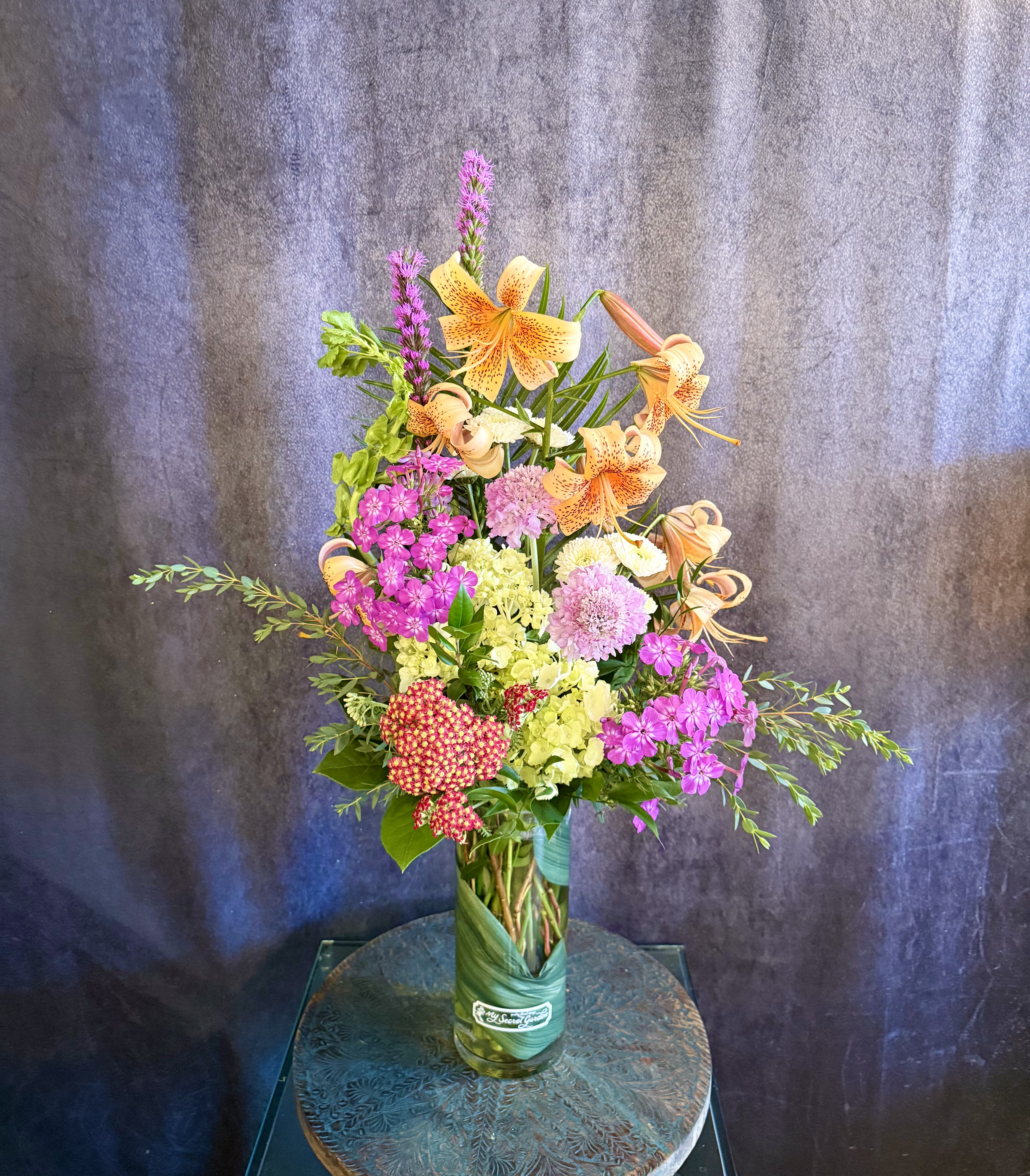 Avant-Garden - The Avant-Garden is a stunning modern design perfect for the person whose creativity shines brightest. Filled with beautiful locally grown flowers from our own Secret Garden such as Tiger Lilies, Red Yarrow, Purple Phlox, and a variety of other seasonal blooms, this arrangement ushers in the summer season with style. Arrangement stands approximately 32in tall and 18in wide.