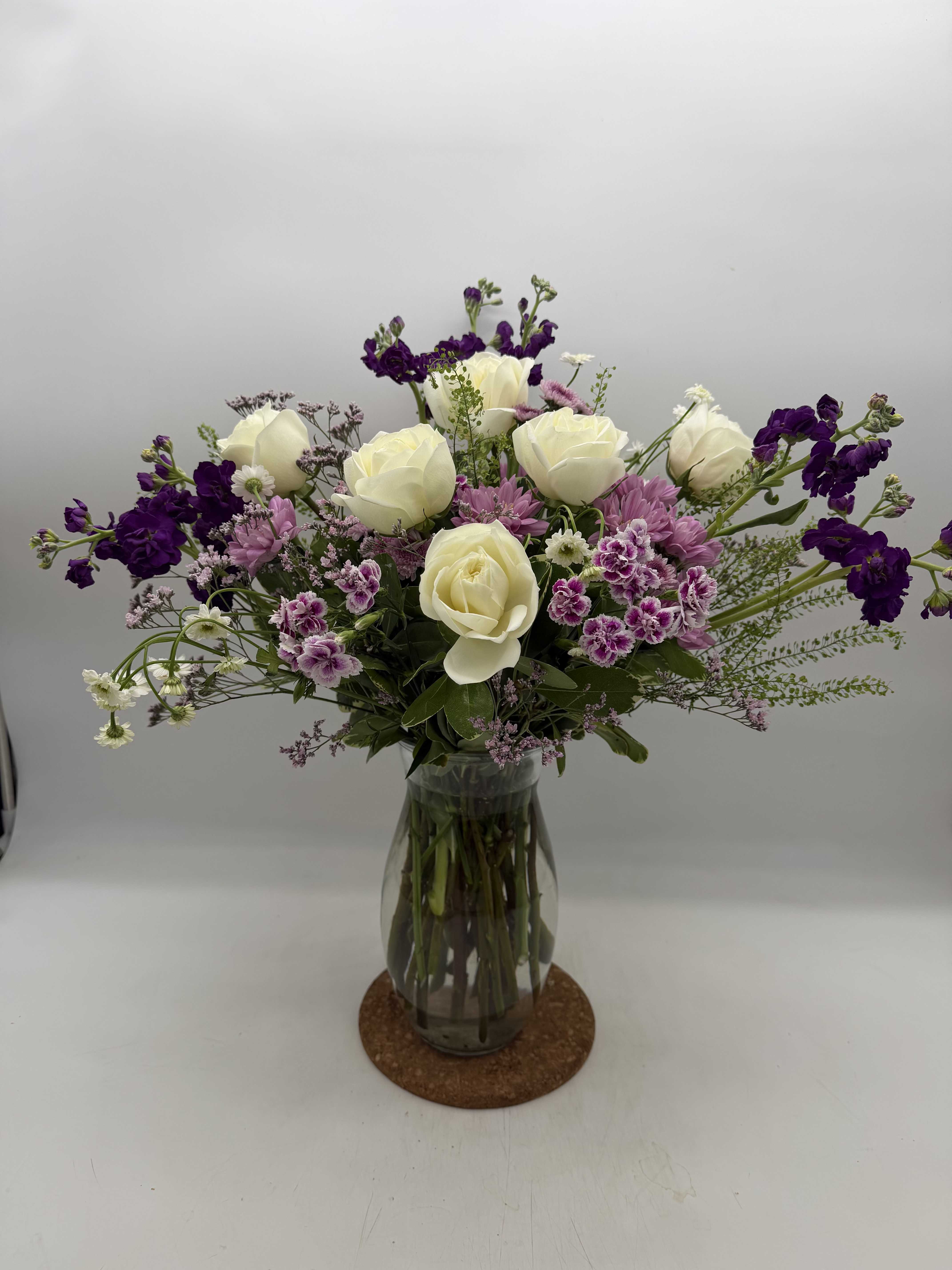 Lush Lavender - Creamy white roses mingle with lavender-hued stock and purple alstroemeria in a lush, rounded vase arrangement that reads like a garden gathered at its most romantic - perfect as a birthday bouquet, anniversary arrangement, or a thoughtful "thinking of you" gift. The layered mix of deep violet tones, soft lavender accents, and touches of pale pink gives the recipient a feeling of being warmly celebrated and truly seen.  This full, dense design is presented in a clear glass vase with visible stems for a fresh, classic look that suits both casual and more formal settings. Expect a compact, rounded silhouette with a rich blend of roses, alstroemeria, stock, and complementary greens that create texture and depth. Available in one vase size, it arrives ready to display; trim stems and refresh the water every few days to keep blooms looking their best.