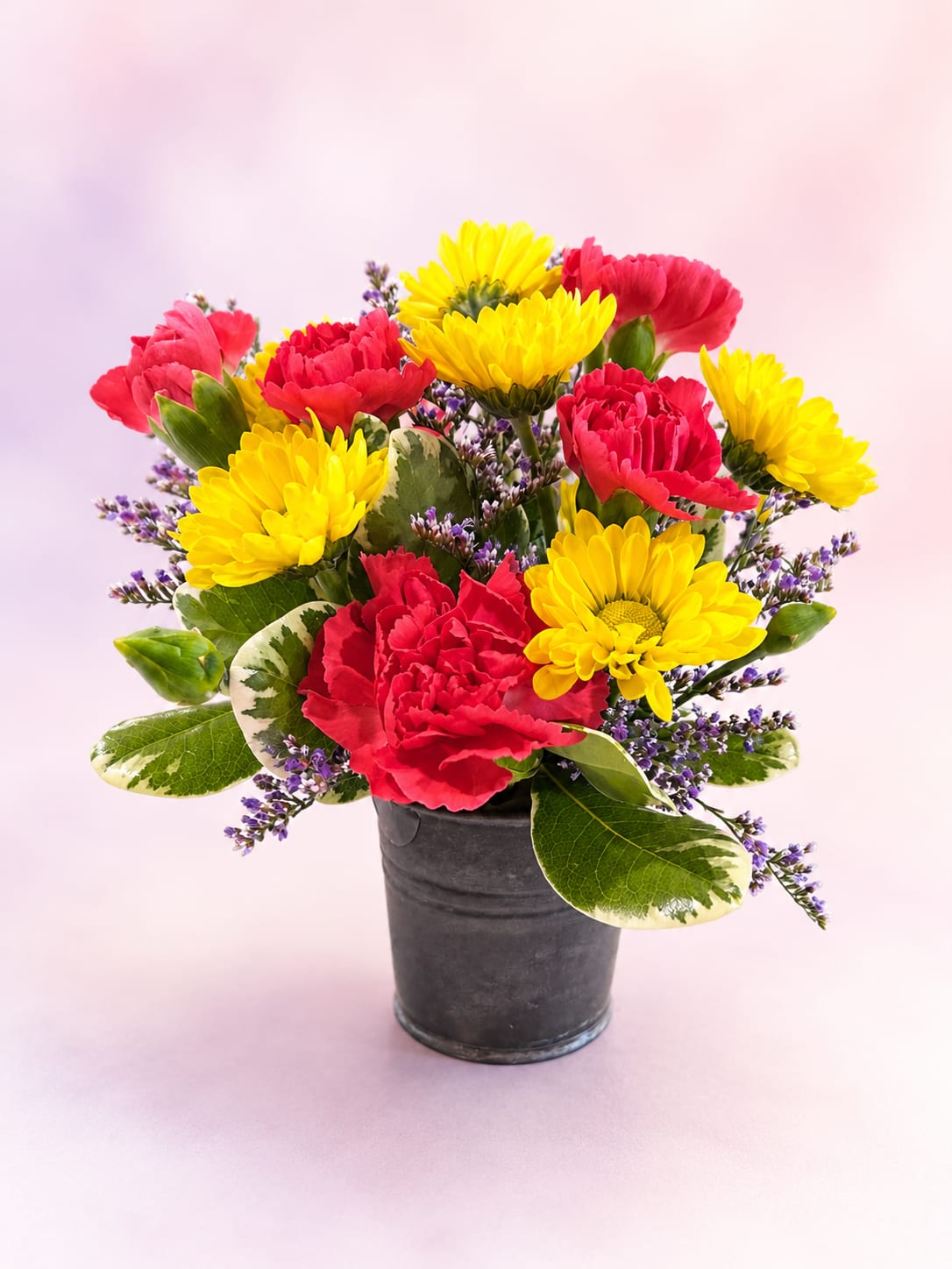 Pink Lemonade - Fresh, vibrant, and bursting with cheerful color, Pink Lemonade is a delightful mini arrangement designed to brighten any day. Featuring bold hot pink mini carnations paired with sunny yellow daisy mums, delicate lavender statice, and variegated greenery, this charming bouquet is arranged in a rustic black metal keepsake container for a touch of farmhouse flair.  Like a glass of sweet pink lemonade on a sunny afternoon, this petite arrangement is full of warmth, happiness, and playful charm. Perfectly sized for desks, countertops, and cozy spaces, Pink Lemonade makes a thoughtful gift for birthdays, thank-you gestures, get well wishes, or simply to let someone know you're thinking of them.