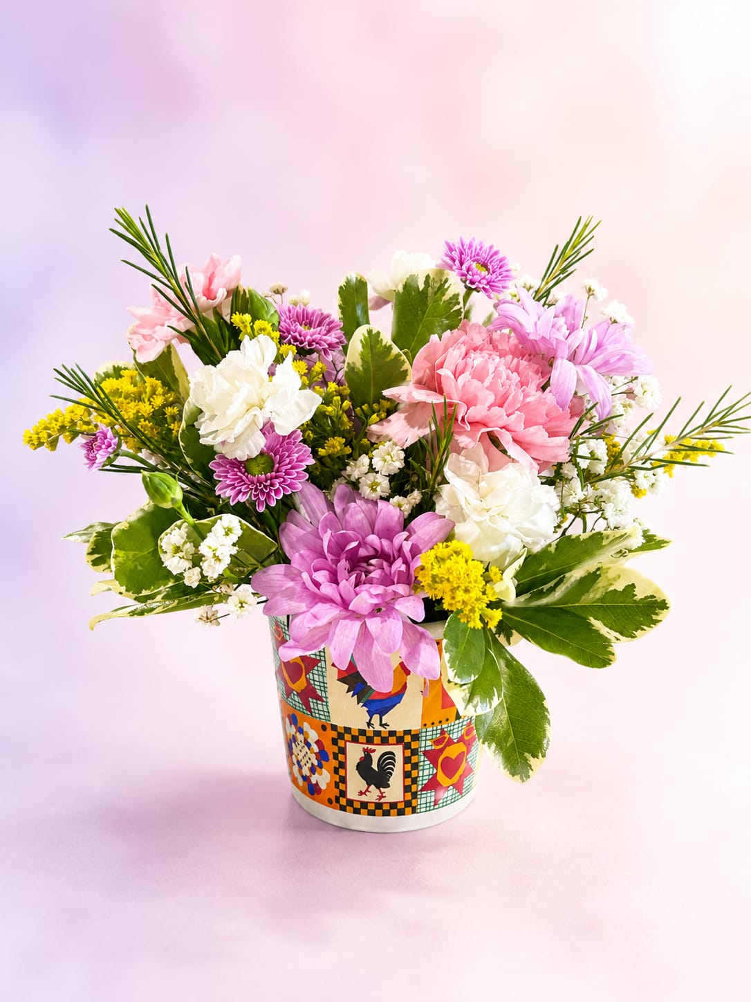 Country Cottage Charm - Sweet, cheerful, and full of homespun beauty, Country Cottage Charm is a delightful mini arrangement designed in a whimsical rooster keepsake container. Featuring soft pink carnations, lavender daisy mums, delicate white blooms, cheerful touches of golden solidago, and lush variegated greenery, this petite bouquet captures the warmth and joy of a cozy country garden.  Overflowing with fresh textures and charming colors, Country Cottage Charm brings a touch of farmhouse elegance to kitchens, desks, and cozy corners alike. Perfect for celebrating birthdays, saying thank you, sending encouragement, or simply brightening someone's day, this sweet arrangement is sure to make anyone smile.