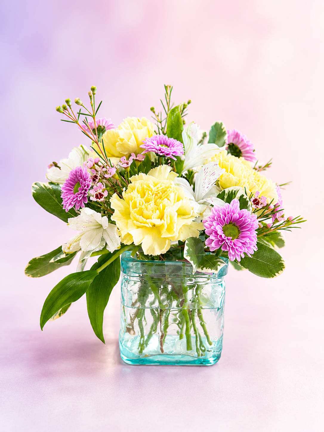 Lemon Lavender Delight - Fresh, cheerful, and effortlessly elegant, Lemon Lavender Delight is a charming mini arrangement that brings together soft colors and graceful textures in a beautiful aqua glass keepsake vase. Creamy yellow carnations mingle with delicate white alstroemeria, playful lavender button mums, and whimsical waxflower accents, all nestled among lush variegated greenery.  With its airy garden-inspired style and soothing pastel palette, this petite bouquet evokes the sweetness of a sunny spring morning. Perfectly sized for desks, bedside tables, and cozy spaces, Lemon Lavender Delight offers a lovely way to celebrate birthdays, send encouragement, express gratitude, or simply brighten someone's day.