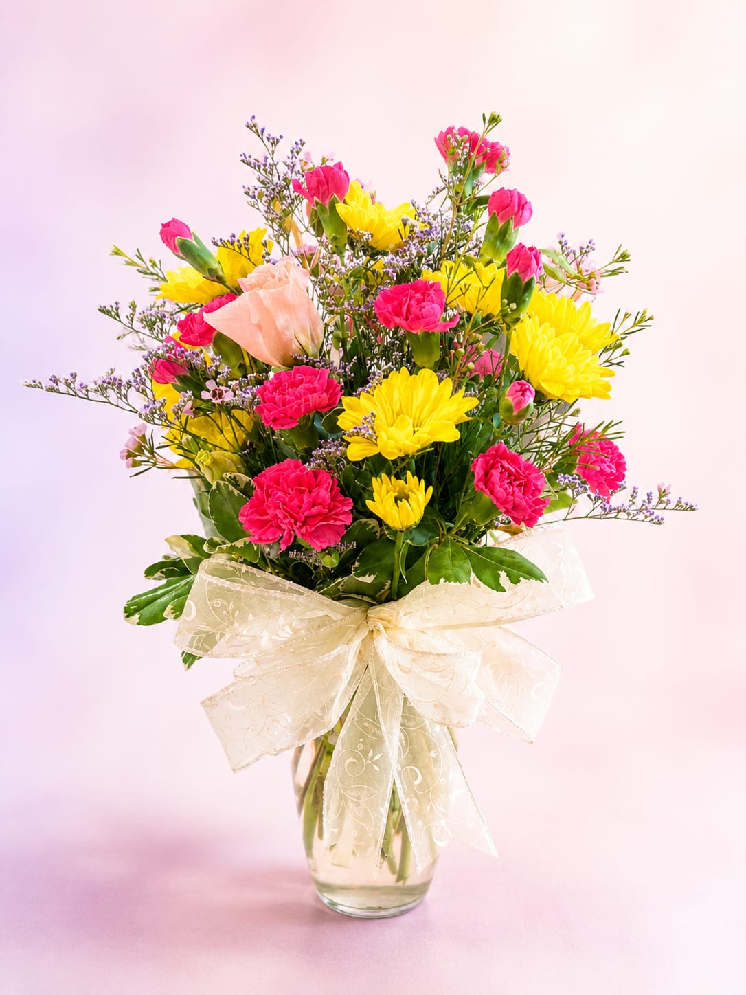 Blush &amp; Bloom - Soft, cheerful, and effortlessly elegant, Blush &amp; Bloom is a graceful arrangement designed to celebrate life’s sweetest moments. Vibrant hot pink carnations and sunny yellow daisy mums are beautifully complemented by delicate blush lisianthus, airy lavender statice, and whimsical waxflower accents, all gathered among lush variegated greenery and finished with an elegant ivory bow.  With its fresh garden-inspired style and charming blend of colors, this bouquet radiates warmth, happiness, and timeless beauty. Whether celebrating a birthday, sending heartfelt thanks, offering encouragement, or simply brightening someone's day, Blush &amp; Bloom is a lovely expression of joy and affection.