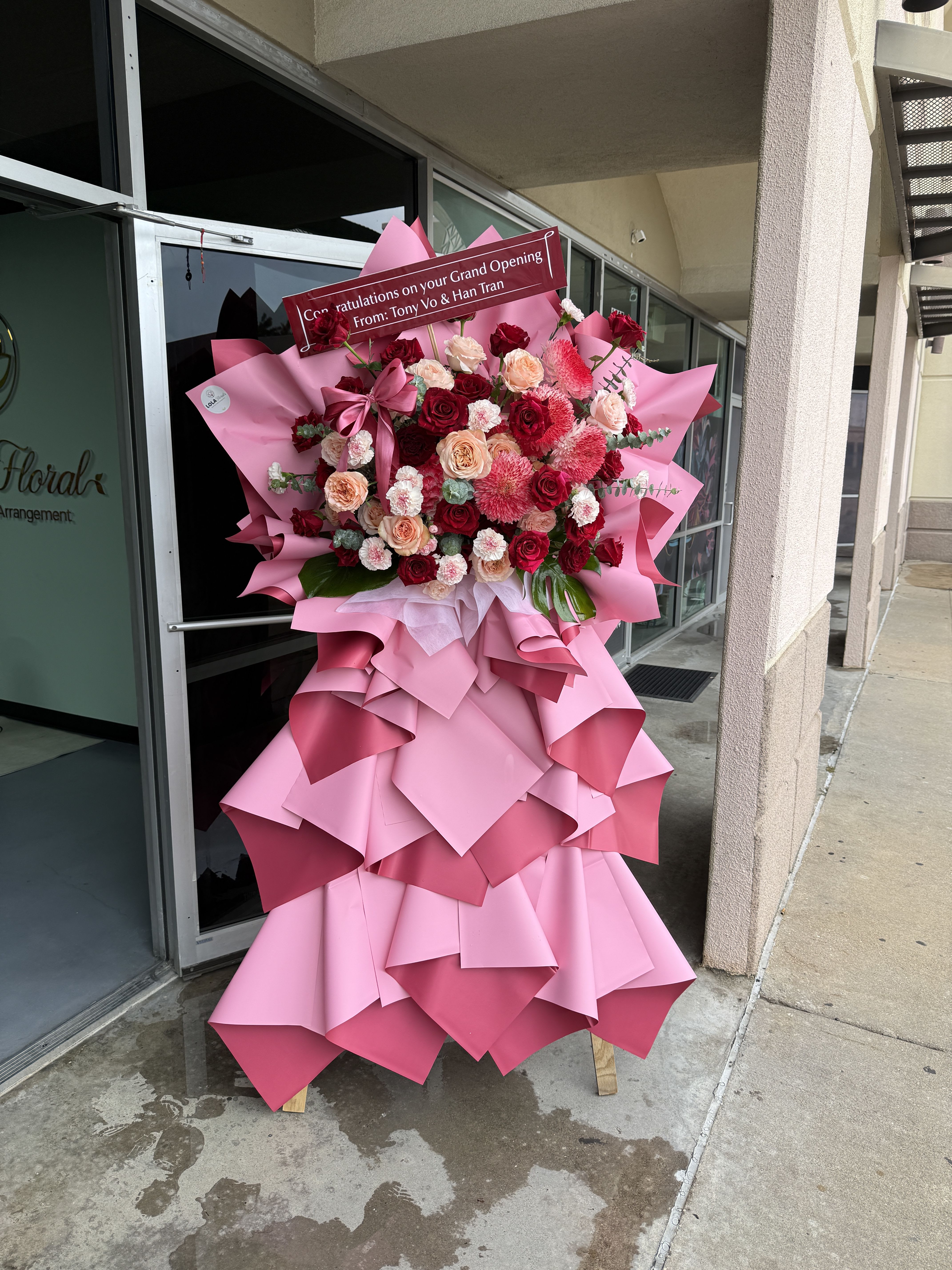 Grand Opening Red 07 - Red, blush, peach and white roses and carnations arranged on a wooden stand with pink wrap, satin ribbon and congratulatory sign-bold, romantic standing display for grand openings.