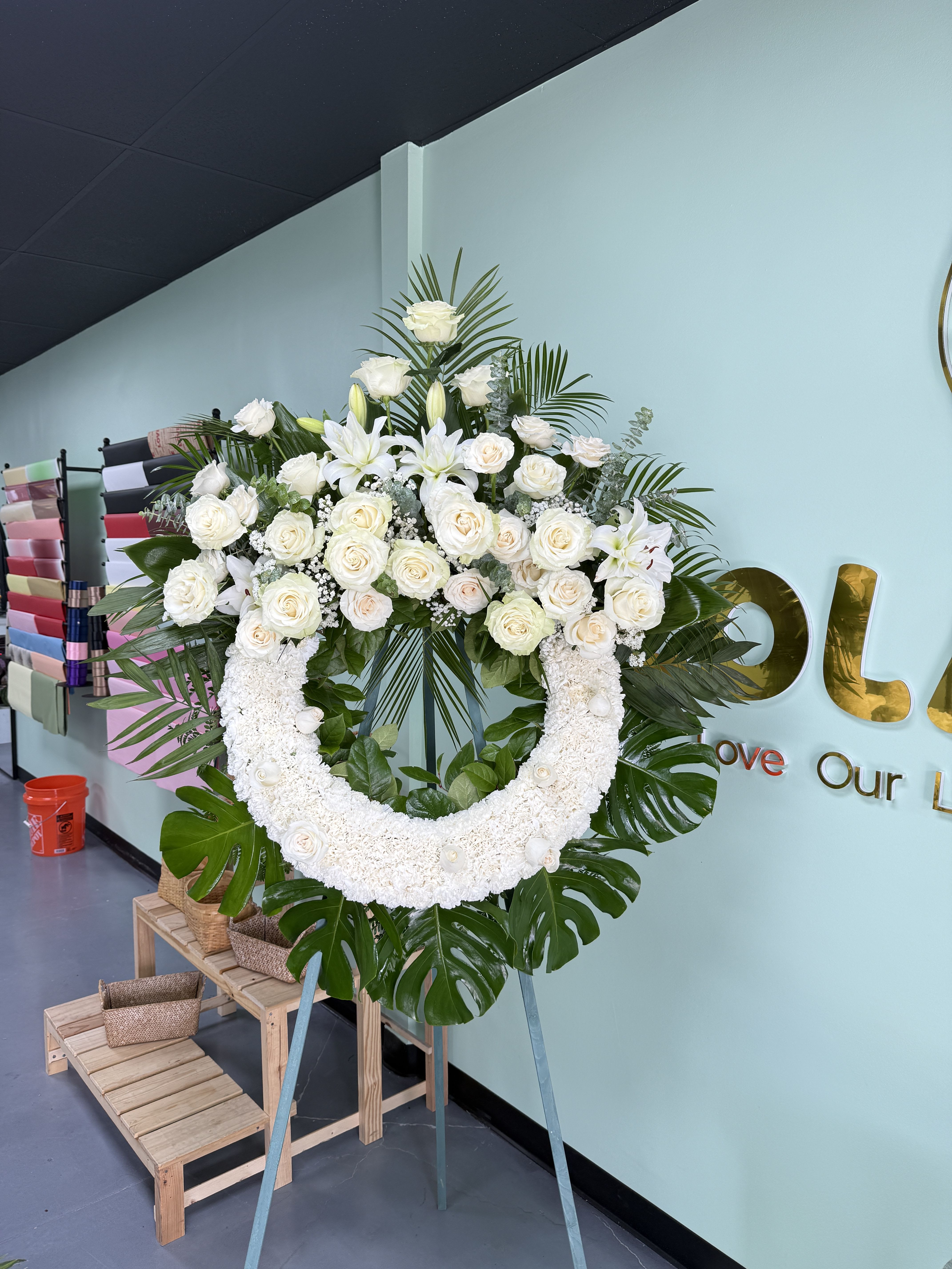 Sympathy Wreath Circle 03 - White chrysanthemums ring with clusters of white roses and lilies atop lush tropical leaves-an elegant, ceremonial standing wreath displayed on a metal easel for sympathy or memorial occasions.