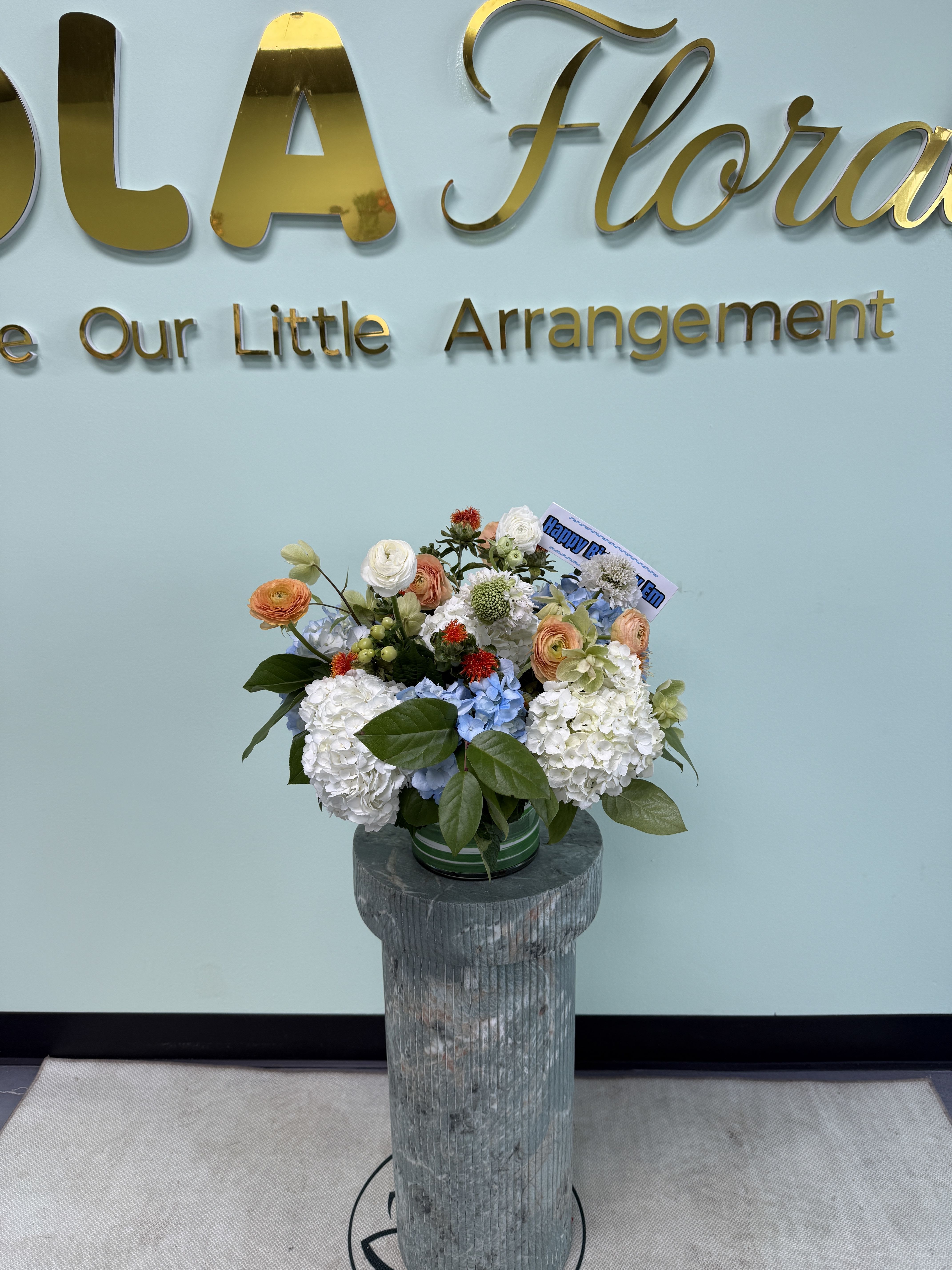 Just Because 01 - Rounded white hydrangeas with pale blue and peach ranunculus, glossy foliage and bright accents in a green container on a decorative pedestal-cheerful everyday gift with a small greeting card.