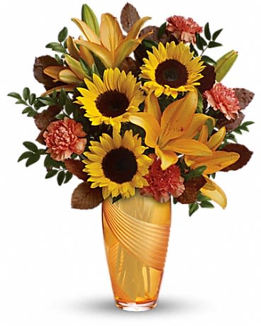 Golden Grace Bouquet - Bright yellow sunflowers and golden-orange lilies give this flower arrangement an immediate sunlit presence-perfect as a summer or spring birthday bouquet or a cheerful Thinking of You gift. The upright, full silhouette in a tall amber glass vase carries warm Thanksgiving and autumnal vibes without feeling strictly seasonal, so she'll feel celebrated whether you're marking a holiday, a thank-you, or just sending a pick-me-up.  The mix pairs bold sunflower heads with coral to red-orange carnations and scattered dark green foliage, plus a few brown accent leaves for a slightly rustic texture. The amber glass vase lifts the palette and lets the stems show through, creating a warm, homey display about the size of a standard tall arrangement-designed to sit on a mantle or dining table and catch the light. The contrast of deep sunflower centers against vivid petals keeps the look energetic and friendly.  Send this to say thanks during Admin Professionals' Week, brighten a Fourth of July gathering with autumnal warmth, or drop it off as an everyday reminder that you're thinking of them. Low-maintenance care: fresh water and a clean vase keep blooms looking their best for days; replace water every other day and trim stems if needed.