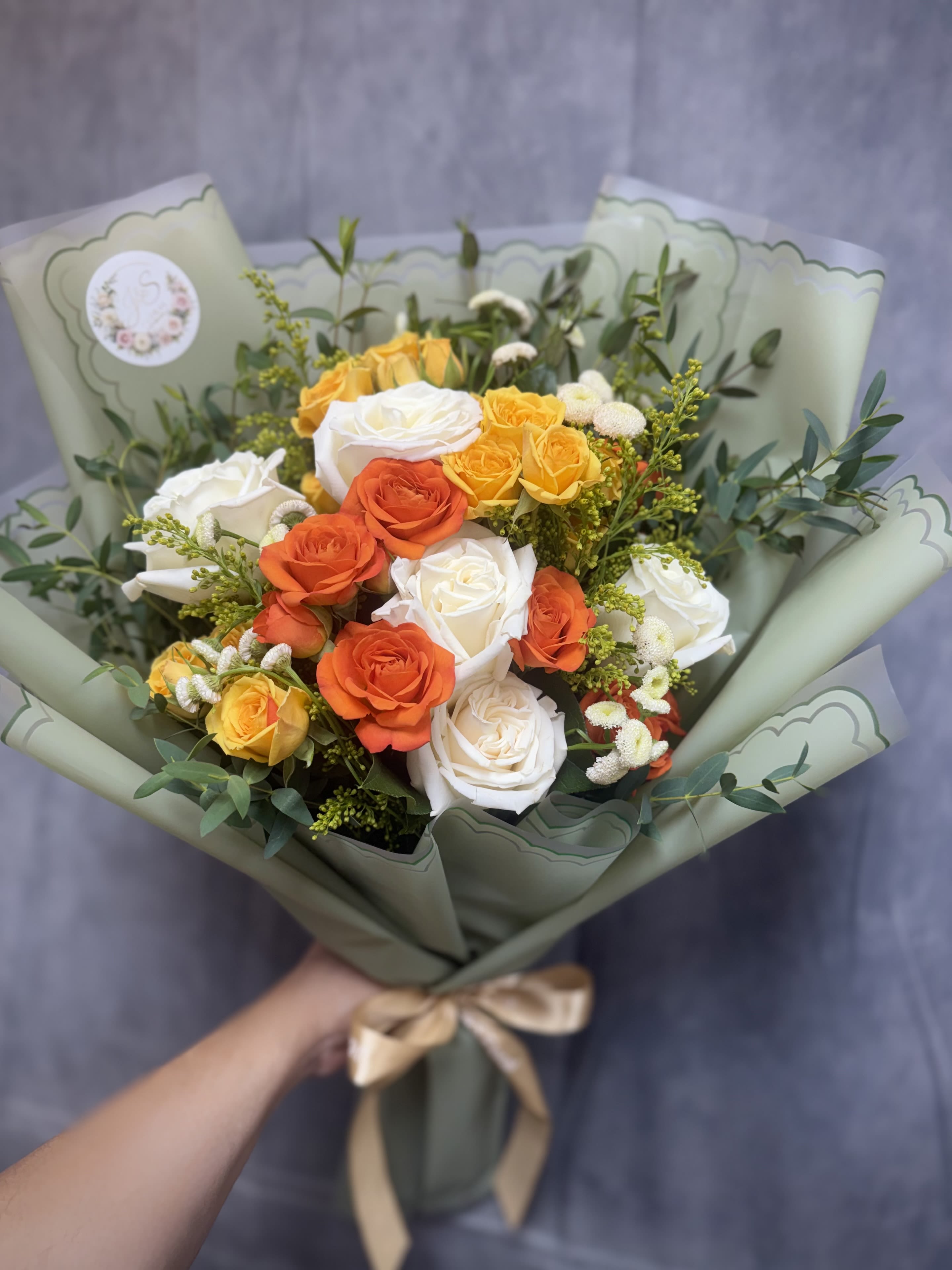 Citrus Meadow - A fresh burst of elegance and sunshine—this hand-tied bouquet features white roses, cheerful yellow and orange spray roses, accented with green waxflower and seasonal fillers. Wrapped in a rich green paper for a refined finish, Citrus Meadow brings the warmth of summer fields to any occasion.