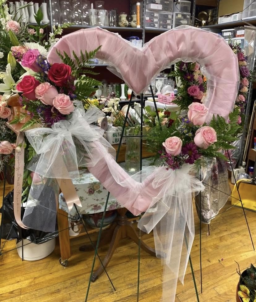 Loving Open Heart on Easel - A large heart-shaped floral wreath on a metal easel stand creates an immediate, heartfelt impression - perfect for a wedding ceremony backdrop or a memorial tribute. The romantic mix of pink and red roses combined with soft white accents makes this a meaningful choice when you want the person you're honoring to feel cherished and remembered.  This arrangement features lush roses in shades of pink and red, accented with small purple blooms and airy white tulle woven through the frame. Pink and satin ribbons drape gracefully across the heart, adding gentle movement and a polished finish. Mounted on a sturdy metal easel, the wreath presents tall and centered for clear visibility at an aisle, altar, or graveside.  Choose this wreath to mark a wedding moment or to pay tribute at a celebration of life - it reads as both tender and formal. The easel-mounted heart arrives ready to display; light misting of the flowers will help preserve freshness on the day of the event.