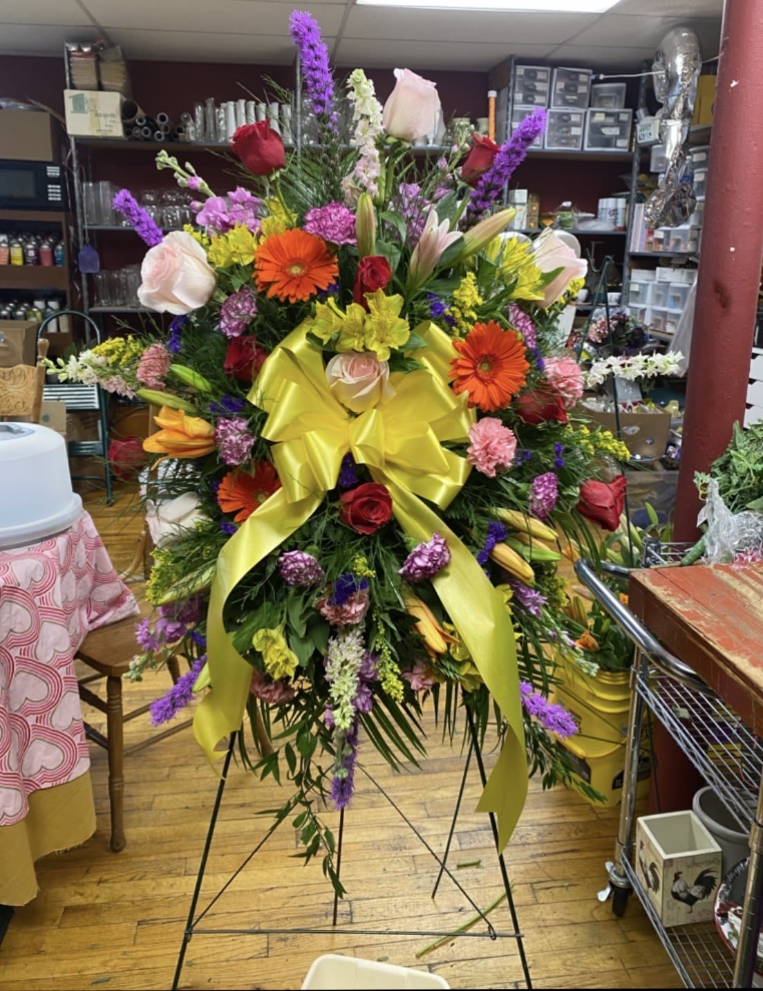 Whimsical Morning Standing Spray - A bold, uplifting sympathy standing spray with high-contrast color-perfect for honoring a life with bright warmth. This Whimsical Morning standing spray combines roses, lilies, gerbera daisies, carnations, snapdragons and alstroemeria in yellow, pink, purple, red and orange tones to create a joyful yet respectful memorial display that reads both celebratory and solemn for funeral or graveside services.  Tall spikes and broad, layered blooms give strong vertical movement and a full, dramatic silhouette on a black easel stand; a bright yellow ribbon tied across the center adds a hopeful, sunny accent. The dense composition is designed to be seen from a distance and to hold up outdoors or in a chapel setting, with mixed textures from the velvety roses and lilies to the crisp snapdragons and cheerful gerbera faces.  Choose this spray when you want the person you're remembering to be celebrated with color-a meaningful way to say you're thinking of their family and their joy. The arrangement arrives ready to display on the included easel.