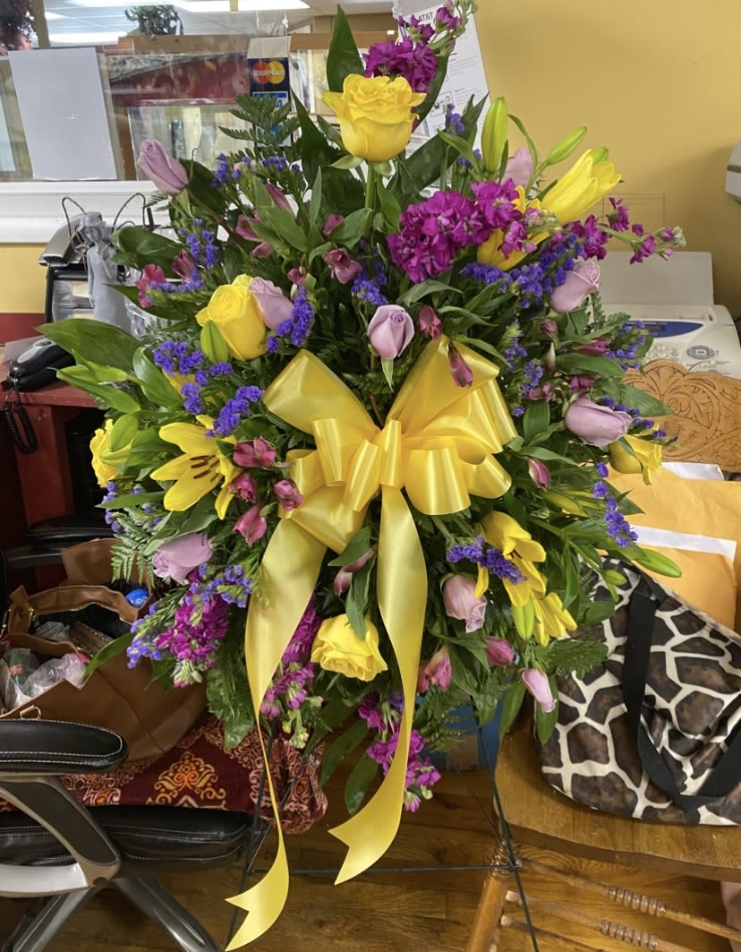 Morning Sunrise Standing Spray  - Sunlit yellow roses and lilies take center stage in this large mixed flower arrangement, perfect as a birthday bouquet, congratulations gift, or bright thank-you surprise. The cheerful yellow ribbon and upright, layered composition announce celebration the moment it's brought into the room-she'll feel genuinely seen and celebrated by its warm, sunny energy.  Dense clusters of yellow roses and lilies are paired with purple and pink accent blooms and glossy green foliage for a lush, abundant look. Alstroemeria and other seasonal flowers add texture and cheerful contrast, while a wide yellow ribbon and floral wrap give a polished, gift-ready finish. The arrangement is designed to make an immediate visual statement and hold its shape as a centerpiece or focal bouquet.  Ideal for sending to a friend, coworker, or family member when you want to lift spirits or mark a happy milestone; it's the kind of bouquet that reads as joyful and thoughtful at once. Standard care-trim stems and refresh the water-will keep the blooms looking fresh, and the substantial size makes it a memorable gift without extra fuss.