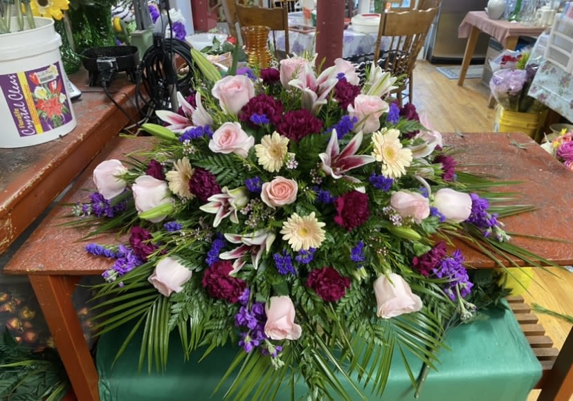 Luscious Purple Casket Spray - A graceful, low-profile casket spray in pink, purple, white, and green creates a comforting, elegant tribute for a funeral or memorial service. This sympathy arrangement features soft blush roses and white lilies at the center, with pale daisy-like blooms and deep violet accents radiating outward-designed to rest gently on a casket or a memorial table and convey tenderness without overwhelming the setting.  The composition is dense and layered, built over mixed greenery and palm fronds for a lush, textured base. Expect classic roses, fragrant lilies, carnations and gerbera-style daisy blooms arranged in a broad, spreading silhouette so the flowers cascade naturally over an edge. The palette of blush, magenta, violet and crisp white offers a romantic, formal feel that reads both respectful and heartfelt.  Choose this spray to honor someone special-whether for a beloved family member, a dear friend, or a colleague-so the person you're remembering will be celebrated with warmth and dignity. Size and placement are intended for casket-top or memorial-table use; contact us for specific dimensions or to request matching standing sprays or smaller sympathy pieces to coordinate with service arrangements.