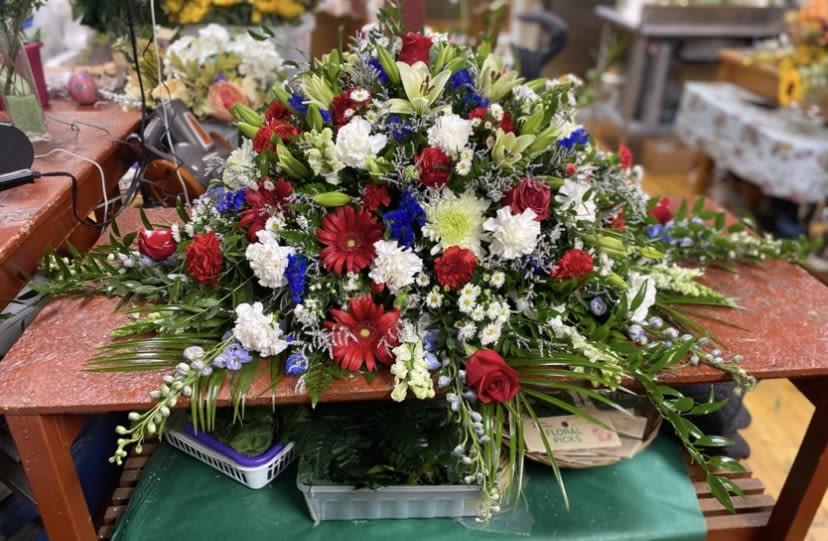 Freedom Casket Spray  - A bold, patriotic casket spray that honors a life with bright, confident color-perfect for Memorial Day, Veterans Day, or any funeral where you want to celebrate service and sacrifice. This freedom-themed flower arrangement reads as a respectful, full-bodied tribute that will make the person you're remembering the focal point of the service.  Dense and low-slung, the spray is packed with red gerbera daisies and red roses set against clusters of white carnations and chrysanthemums, pale lilies, white daisies, and touches of blue accent flowers and alstroemeria. The mix of textures-velvety roses, ruffled carnations, spiky chrysanthemum blooms and soft lily petals-creates a layered, abundant shape in classic red, white, and blue with fresh green foliage. Its substantial size and sprawling silhouette make it suitable for placement on a casket or as a large presentation piece at a memorial service.  Designed to convey honor and remembrance, this arrangement will help the bereaved feel seen and comforted during a difficult moment. As a finished spray, it arrives ready to place on display; standard florist care-keep blooms cool and out of direct sun-will help the flowers look their best through the service.