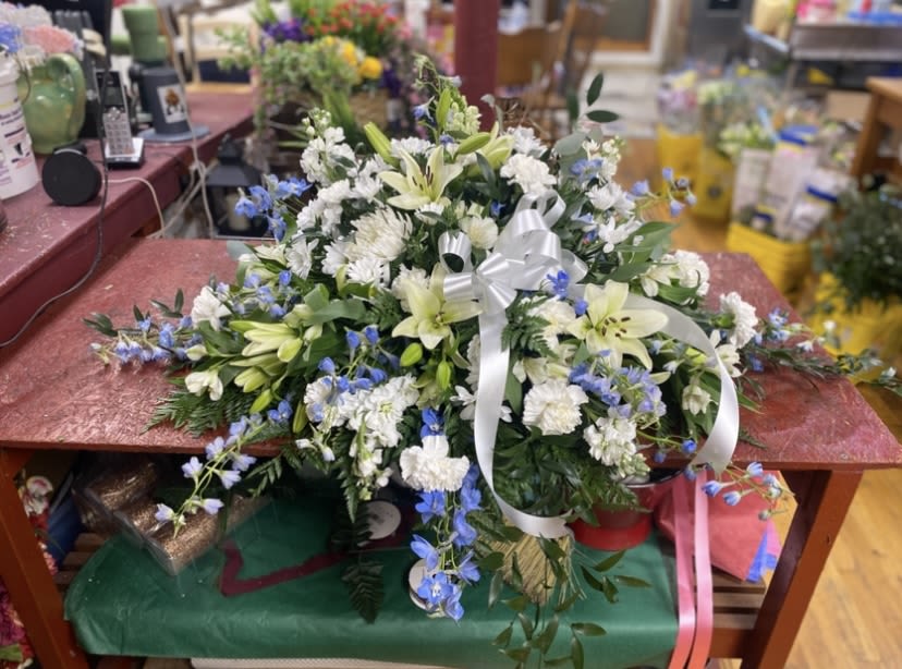 Gracious Light Casket Spray - Soft white lilies and lush chrysanthemums form a serene, full casket spray designed to comfort and honor - ideal for sympathy tributes and moments of quiet remembrance. The arrangement's calming palette of white, pale blue delphinium accents, and gentle green lily buds creates a layered, outward-spreading composition that reads as both formal and understated, making it a respectful choice for funerals or memorial services.  This low, expansive design sits close to the surface and is finished with a white ribbon bow near the center for a polished touch. Textural contrasts between the smooth lily buds, the rounded chrysanthemum heads, and the spiky delphinium stems give the spray depth and movement while maintaining a tranquil, cohesive look suitable for viewing from all sides.  Made to convey sympathy and thoughtful remembrance, this spray offers a dignified way to say you're thinking of someone or to honor a life well lived. Its spread-style shape and classic color story make placement straightforward at services; minimal care is required beyond standard floral water source handling prior to delivery or placement.