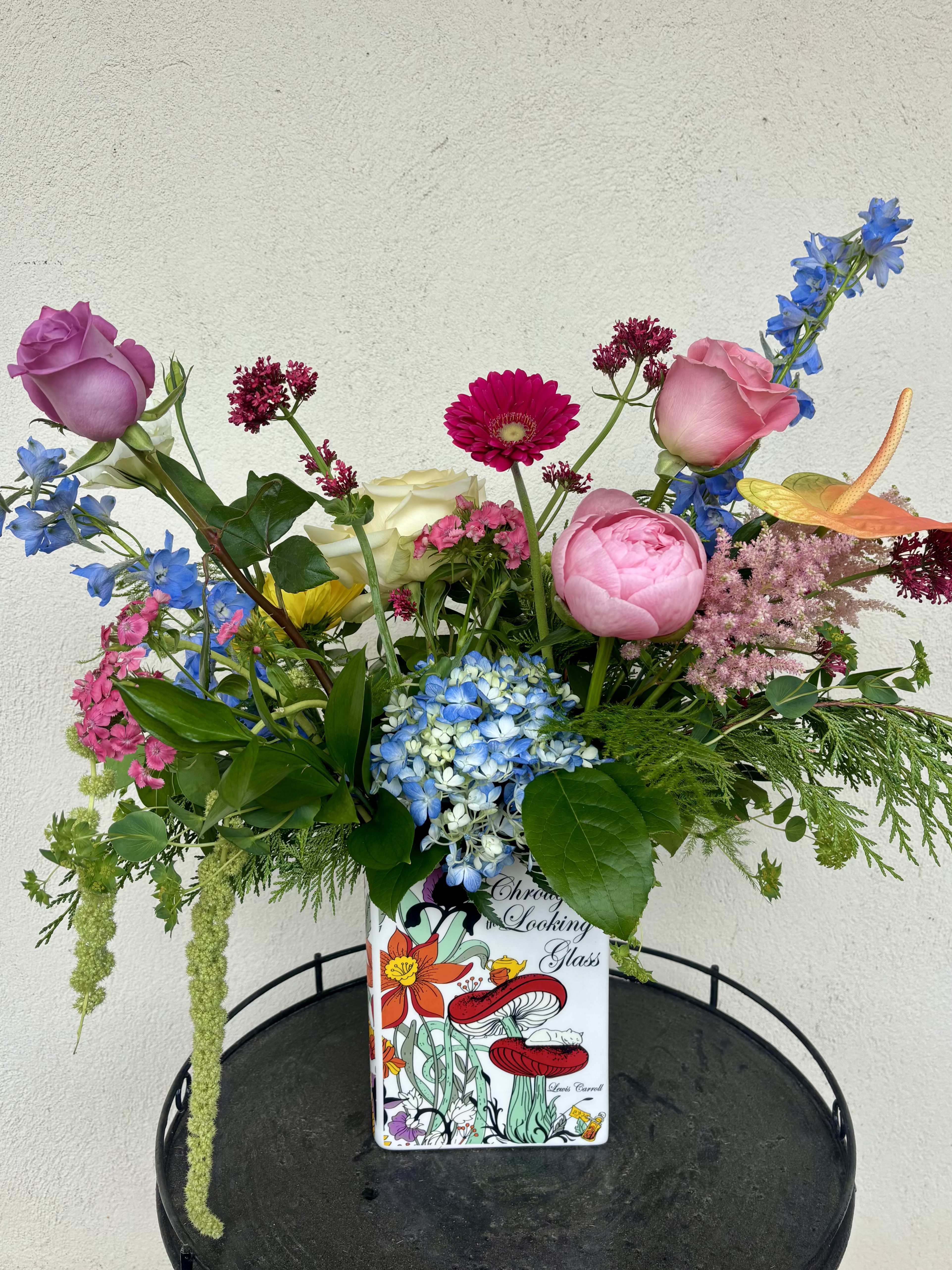 Through the Looking Glass - ***Limited edition and one of a kind arrangement.   Step into a world where imagination blooms. Inspired by the whimsical charm of Through the Looking Glass, this colorful, garden-style arrangement features premium seasonal flowers in a playful palette of vibrant hues and enchanting textures. Overflowing with storybook charm and lush garden beauty, it's the perfect gift for book lovers, dreamers, and anyone who believes a little wonder belongs in every day.