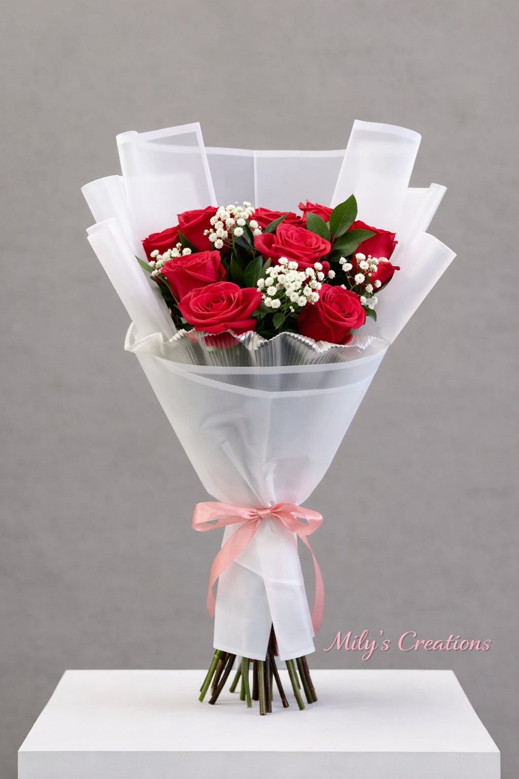 Bouquet of red roses with small white filler flowers wrapped in white paper and tied with a pink ribbon.