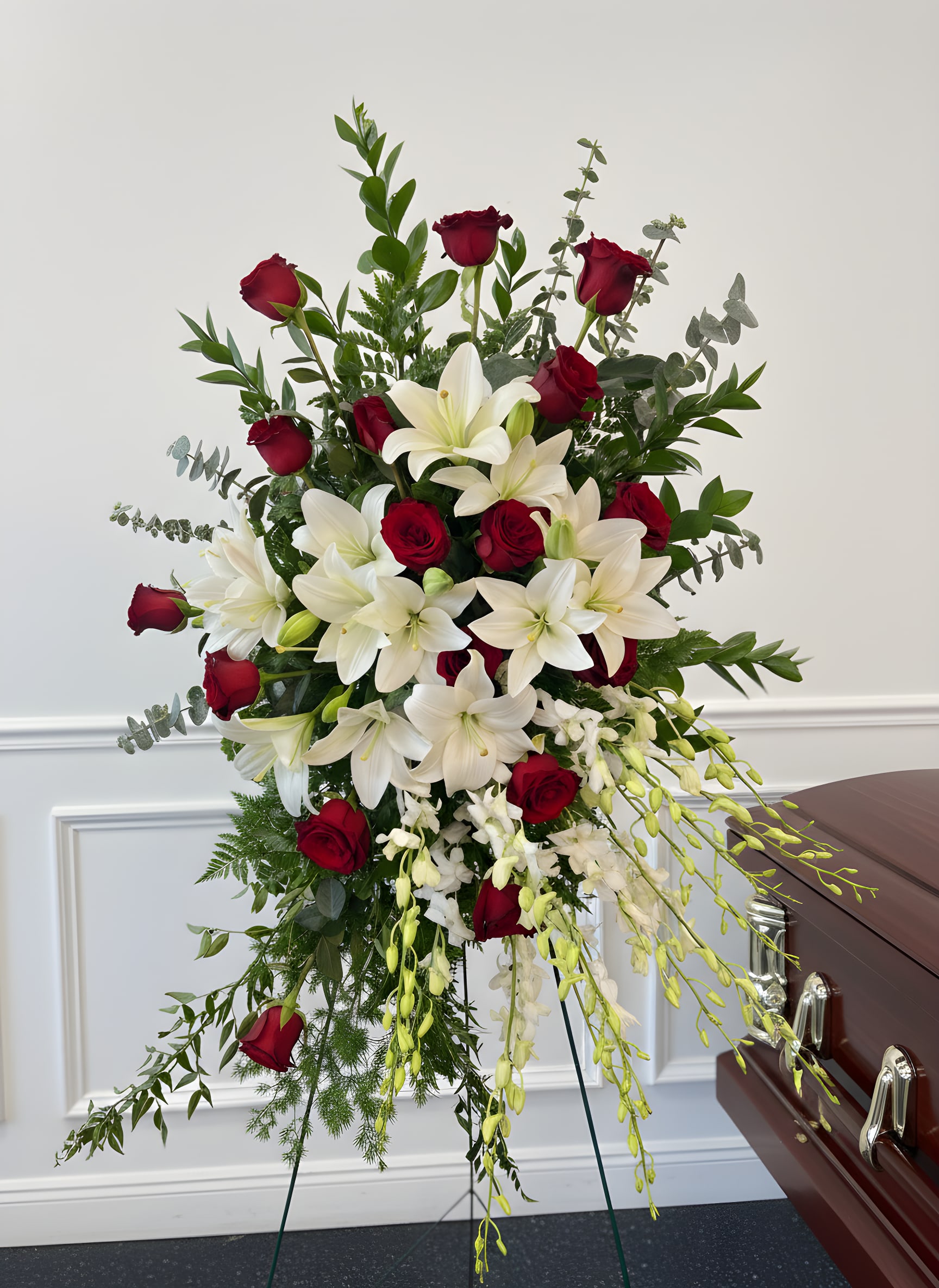 Standing funeral spray with red roses, white lilies, and trailing orchids beside a casket