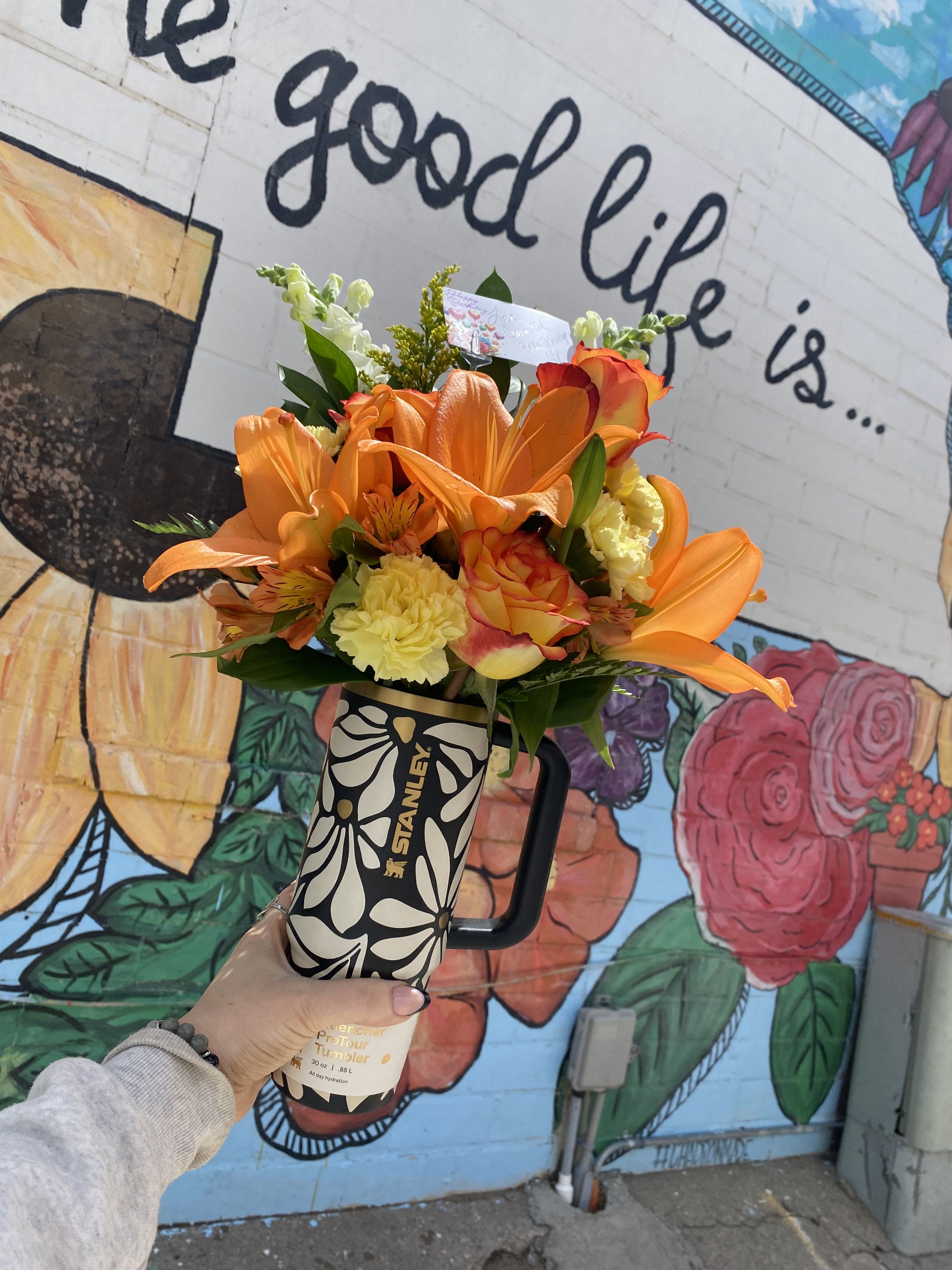 Meadow Morning - Bright orange lilies take center stage in this cheerful birthday bouquet designed to arrive ready-to-gift. The bouquet pairs those showy lilies with sunny yellow carnations, orange-and-pink spray roses, alstroemeria, and crisp white accents to create a warm, energetic palette perfect for birthdays, thank-you gestures, or a just-because pick-me-up. Presented in a black Stanley travel tumbler instead of a traditional vase, it feels modern and practical-part bouquet, part gift set.  The arrangement is gathered compactly into the tumbler so the blooms sit close together for a lush, full look with plenty of texture from ruffled carnations, layered rose petals, and the lilies' dramatic trumpet shapes. The travel tumbler adds everyday usefulness and includes a gift tag for a personal message. This is the kind of gift that makes the person you're thinking of feel seen and celebrated-bright, long-lasting color and a handy keepsake they can use long after the flowers are gone.